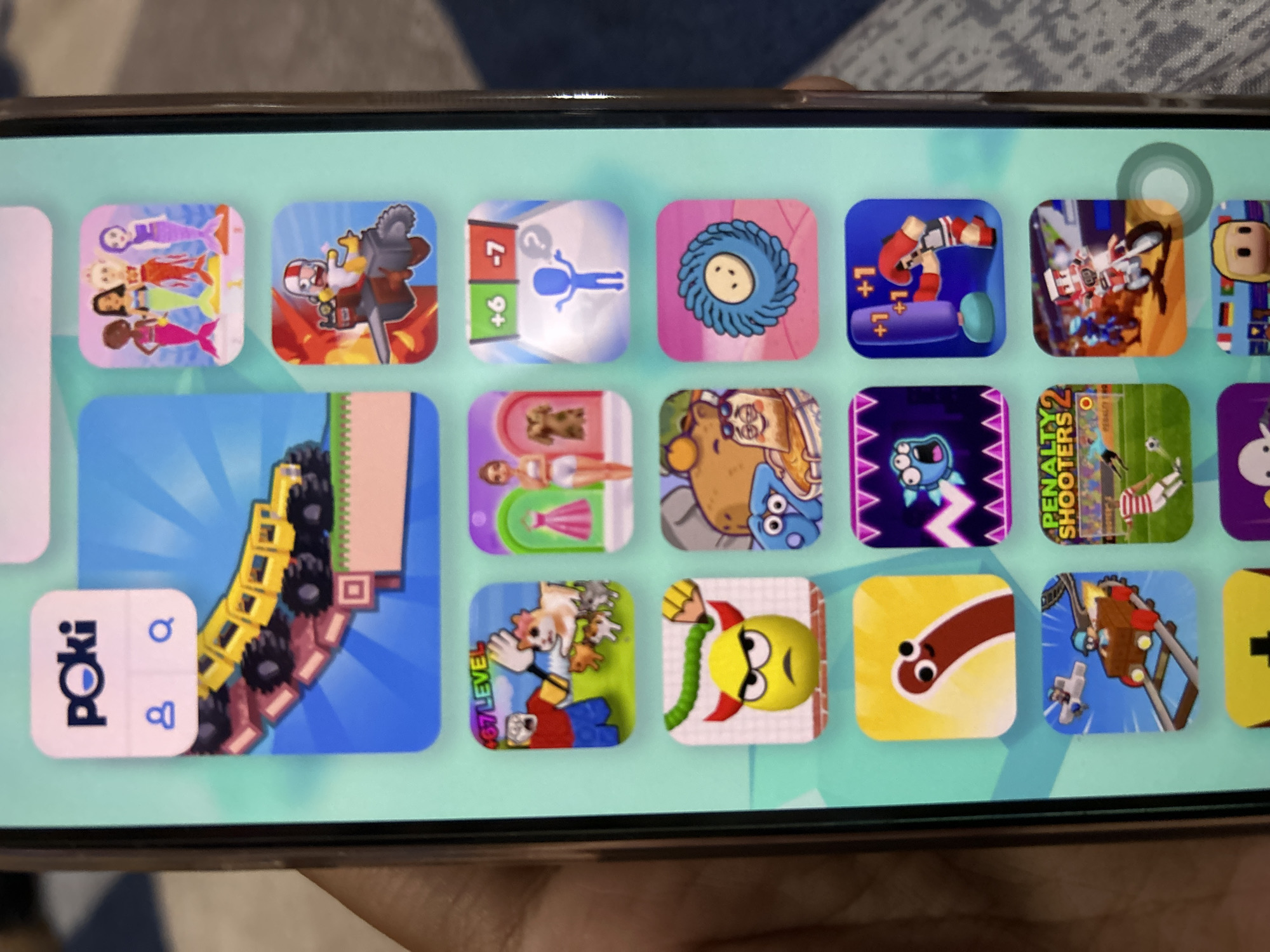 The image shows a smartphone screen displaying a collection of mobile game icons. The screen is held in a person's left hand, with part of their thumb visible on the left side of the frame. The icons are arranged in a grid and represent various games with colorful and cartoonish graphics. Some of the visible game icons include a yellow monster truck, mermaids, a person boxing a punching bag, a worm-like character, a roller coaster, and a soccer game. The overall aesthetic suggests a collection of casual or hyper-casual mobile games. The image was taken in the city of Depok, Indonesia.
