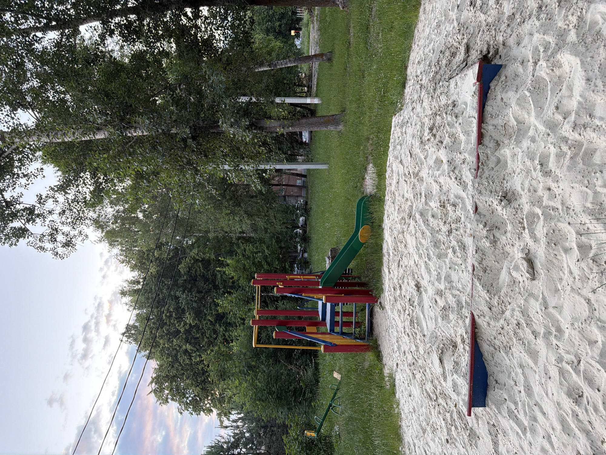A bright, overcast day at a public playground in an urban setting. The foreground is dominated by a large, uneven mound of light-colored sand. Embedded in the sand are three low, rectangular platforms with red tops and blue undersides, arranged in a diagonal line across the mound. To the left of the sand mound, partially in the sand and partially in the grass, is a small green seesaw with two wooden seats. Behind the sand mound is a colorful playground structure with red, yellow, and blue poles and stairs, a blue platform, and a green slide. The playground is situated on a patch of green grass. Beyond the playground, a dense line of lush green trees forms the background, with a few taller birch trees with white bark visible among them. In the distance, through the trees, are glimpses of apartment buildings and parked cars, indicating an urban environment. Power lines stretch across the sky in the upper left portion of the image. The sky is a pale blue with scattered, wispy white and grey clouds.