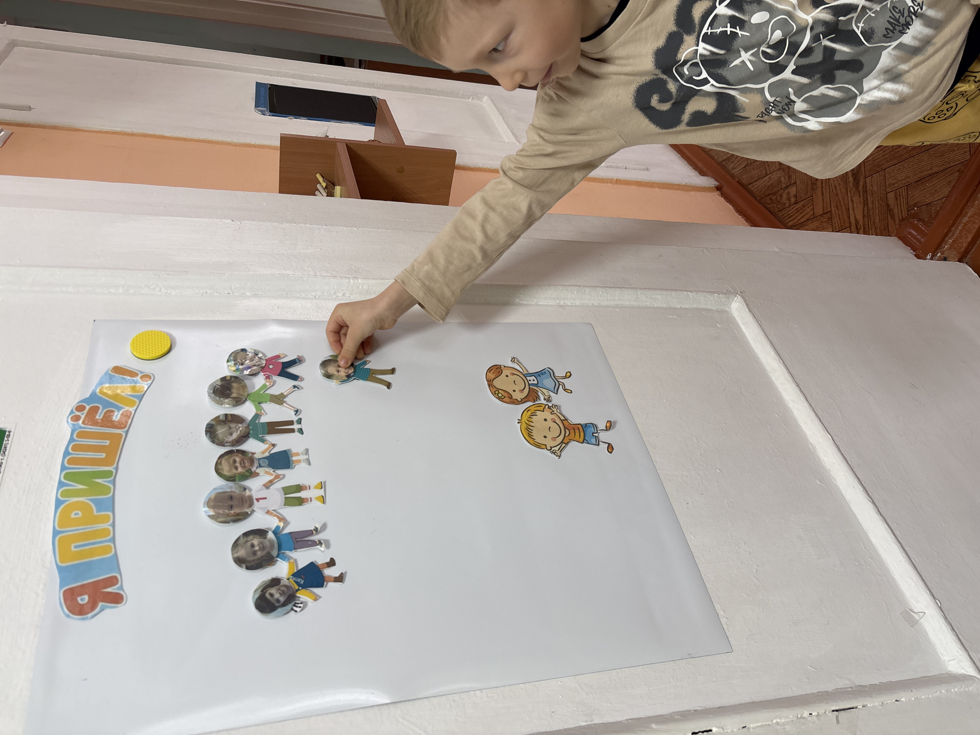 A child with blond hair, wearing a beige long-sleeved shirt with a bear design, is attaching a cutout of a person to a white poster. The poster is mounted on a white wall and features a group of similar cutouts arranged in a vertical line, as well as two separate cutouts to the right. A yellow circular object is at the top left corner of the poster, and text in blue and orange is present next to it. Above the poster is a peach-colored wall section with a small wooden shelf holding items, and above that is a white surface. The floor appears to be covered with brown tiles. The image was taken in the city of Chaikovsky, Russia.