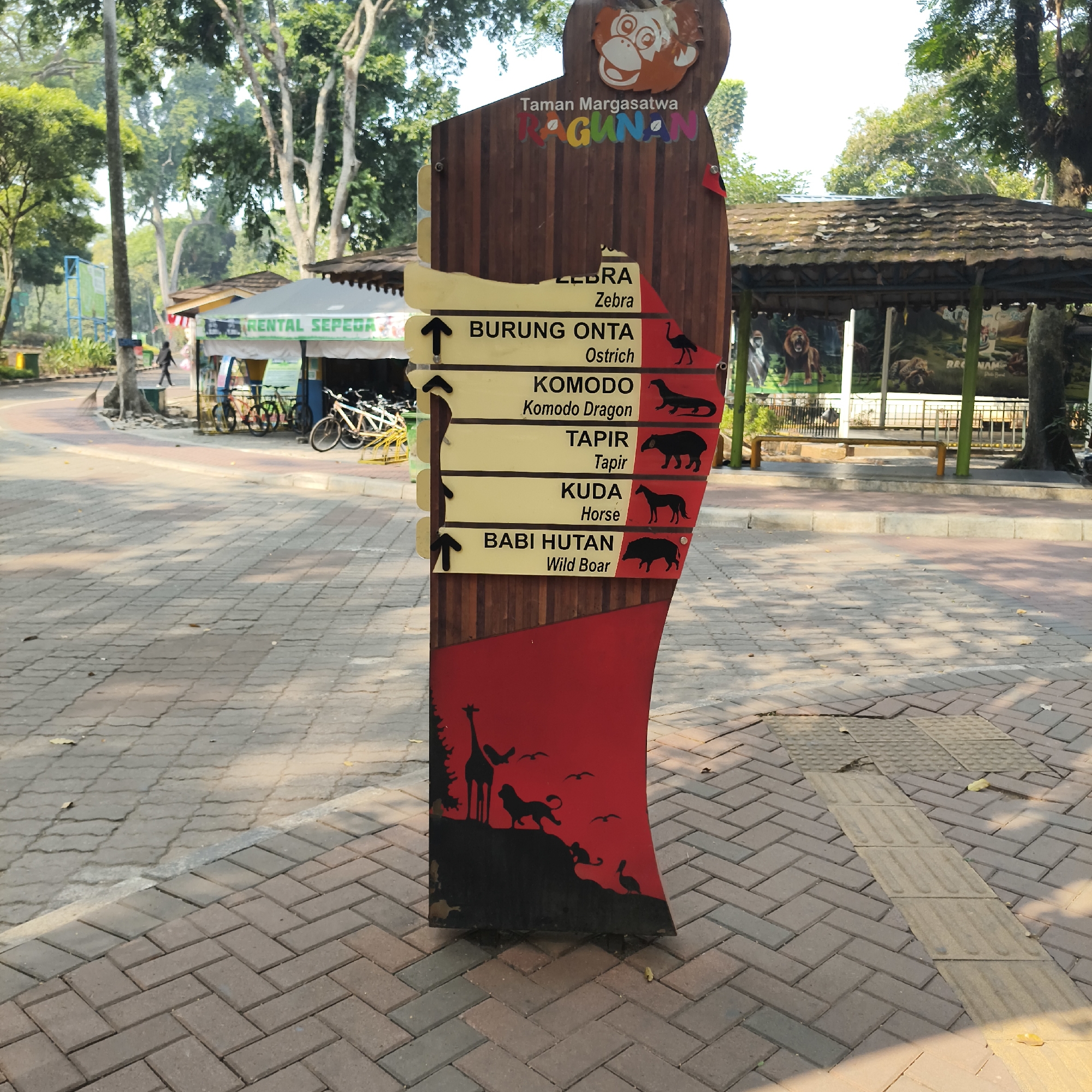 The image shows a directional signpost in Taman Margasatwa Ragunan, a zoo located in South Jakarta, Indonesia. The signpost is made of wood and features several signs pointing in different directions, indicating various animal exhibits. The signs are labeled in Indonesian and English, with animal names such as "ZEBRA," "BURUNG ONTA" (Ostrich), "KOMODO" (Komodo Dragon), "TAPIR" (Tapir), "KUDA" (Horse), and "BABI HUTAN" (Wild Boar). Each sign is accompanied by a silhouette of the corresponding animal. At the base of the signpost, there is a red section with silhouetted images of a giraffe, a lion, a monkey, and birds.
In the background, there are trees, a building with a tiled roof, and a rental bicycle stand with several bicycles. The ground is paved with brick tiles. The lighting suggests it is daytime, with shadows cast by the trees and the signpost. No people are prominently featured, though one person is visible walking in the distance near the bicycle rental.