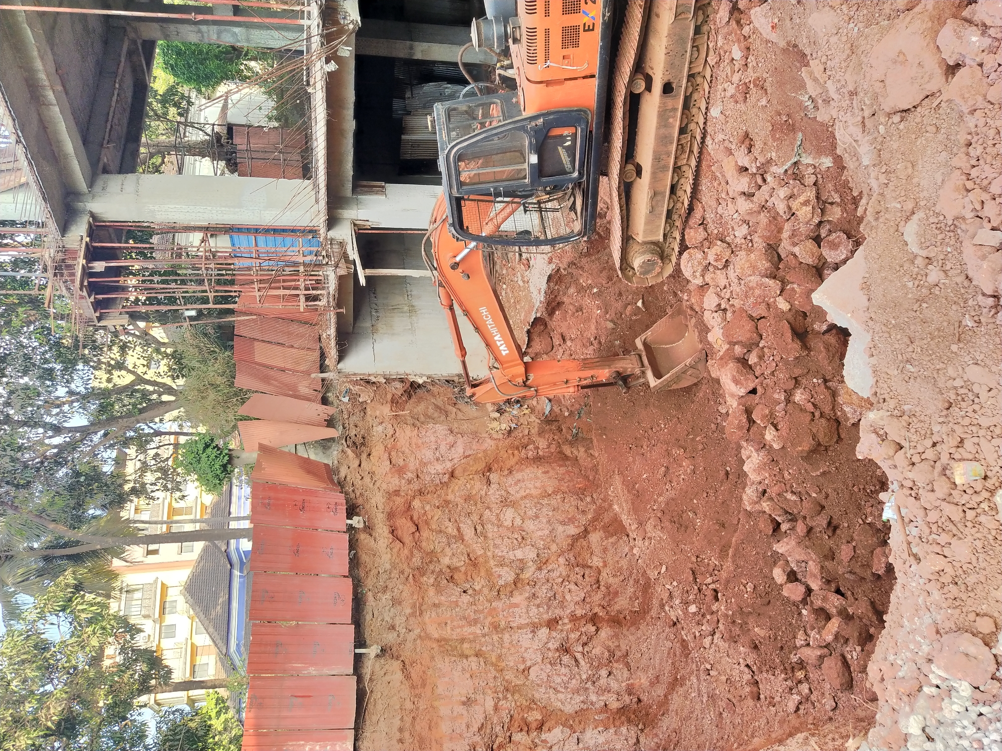 An excavator digs a deep trench at a construction site in Mapusa, India. The construction site is located adjacent to a building with a red retaining wall and a building with a blue tiled roof. The excavator is orange and black, and the excavated dirt is red. There are no people visible in the image.