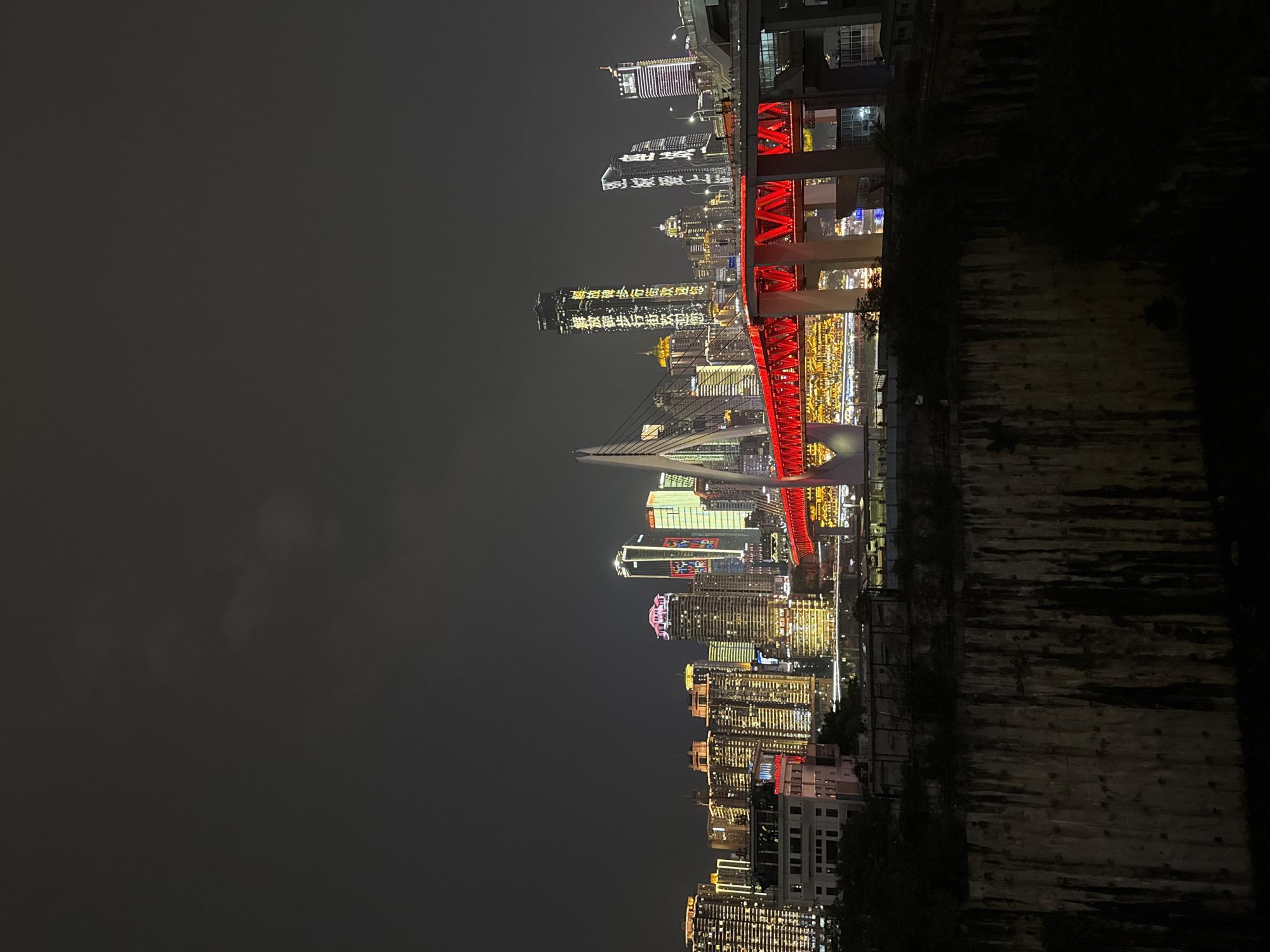 This is a nighttime, low-angle shot of a cityscape in Jiangbei Cheng Subdistrict, China. The image is oriented vertically, showing a dense cluster of illuminated skyscrapers and a prominent red bridge. The bridge, a focal point, features a brightly lit, arched structure with red accents and supports. Behind the bridge, a multitude of buildings of varying heights are lit with a mix of white, yellow, and some red lights, creating a vibrant urban scene against the dark, cloudy sky.
The time of day is clearly night. The weather appears to be overcast, with the sky being a uniform dark gray. The lights of the city reflect faintly on a dark, textured surface in the foreground, possibly a body of water or a wet embankment, suggesting the city is situated near a river. The architecture suggests a modern, bustling metropolis.
There is visible text on the buildings. One building in the middle ground has characters illuminated in yellow that appear to be Chinese script. Another skyscraper to its left has "SWISSÔTEL" and "CHONGQING" written in white lights. Further to the left, a building displays "HOTEL" in white, followed by a logo. The overall impression is one of urban dynamism and scale.