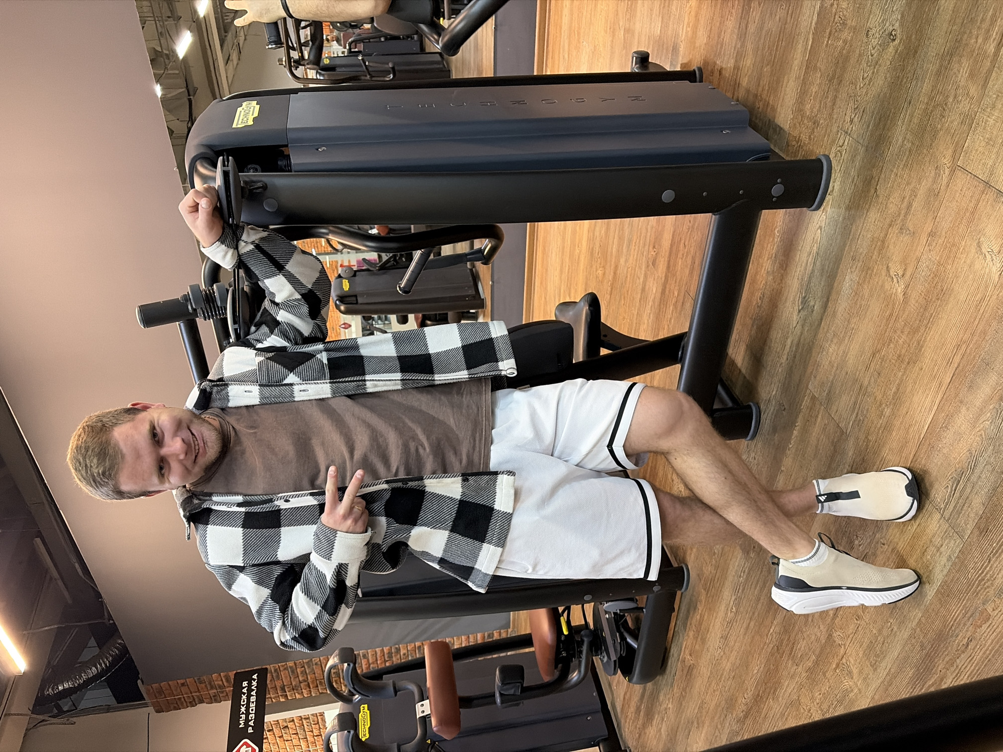 A man is sitting on a weight machine in a gym. The man is fair-skinned with short, light-colored hair, wearing a brown t-shirt, white shorts with black trim, a black and white checkered flannel shirt, and tan sneakers with white soles. He is making a peace sign with his right hand and smiling at the camera. He appears to be seated in a Technogym brand weight machine. The gym has wooden flooring and appears to be well-lit. There is a sign partially visible in the lower left corner of the frame, which includes the text "МУЖСКАЯ РАЗДЕВАЛКА" (Men's Dressing Room in Russian) and the city of Chaikovsky, Russia can be inferred from the writing on the sign.