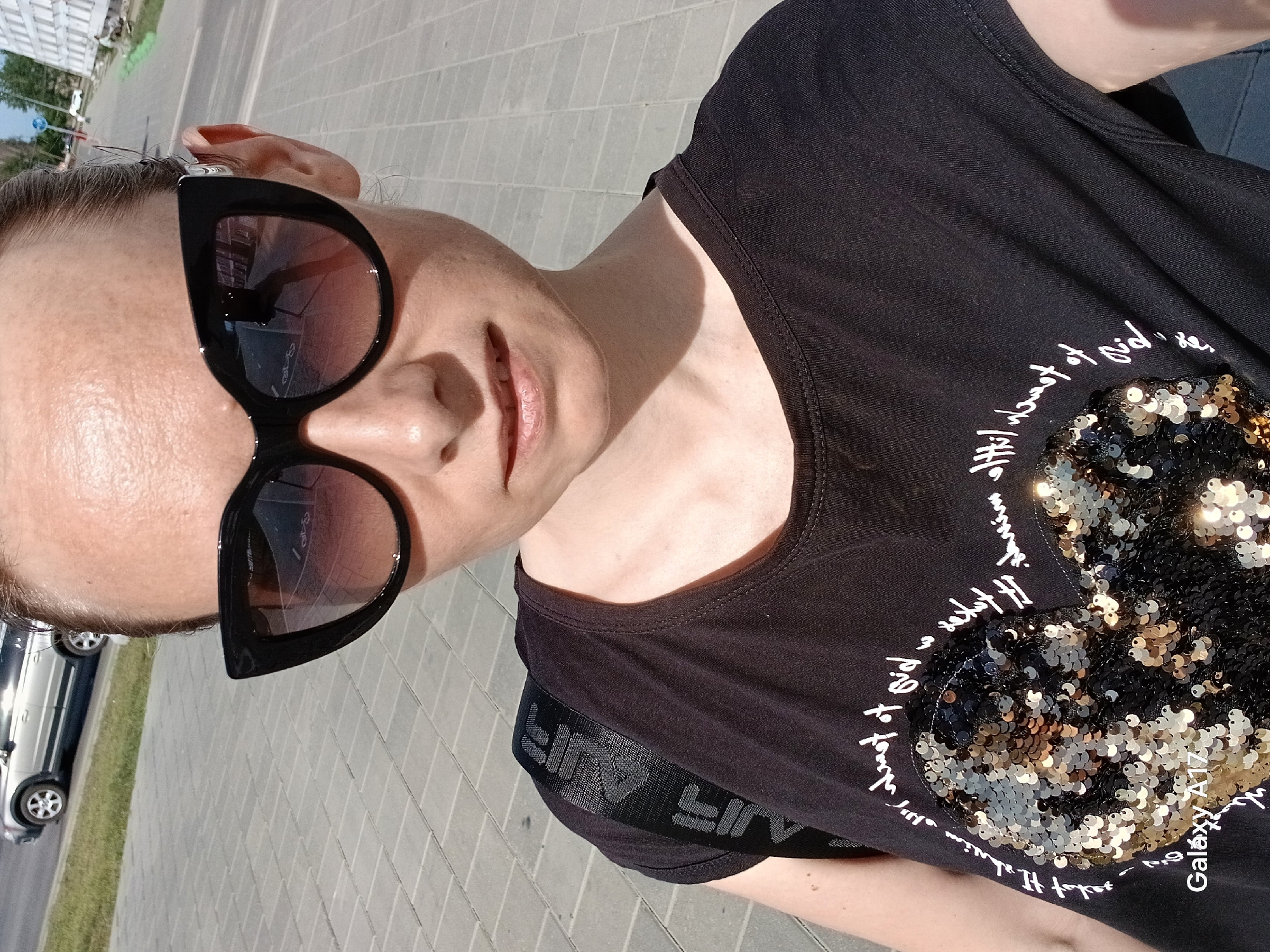 A light-skinned woman, wearing sunglasses and a black t-shirt with sequins, is outdoors. The location is suggested by the vehicle visible in the background and the brick paved sidewalk, implying an urban setting. The perspective suggests the photo was taken at a low angle or selfie-style.