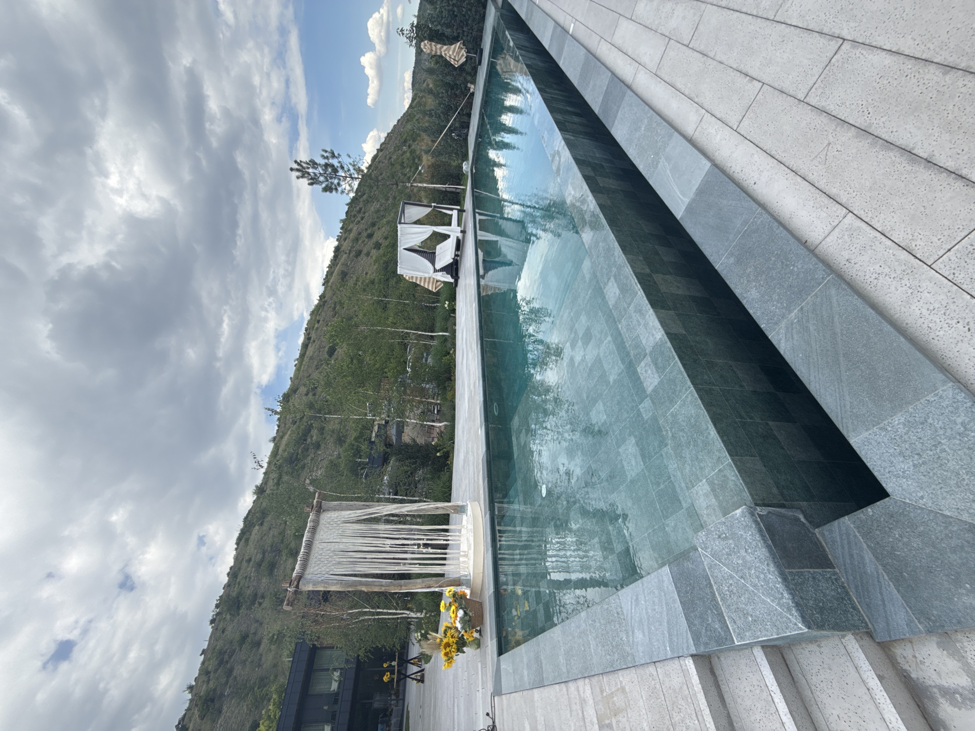 A wide, angled shot captures a modern infinity-edge swimming pool in Morovaia, Moldova. The pool's tiling is a mosaic of dark green squares. The pool is bordered by a stone deck on the right and a grey stone patio with stairs on the left. In the background, a lush green hillside rises, dotted with trees. On the left side of the image, a modern building with large windows is partially visible. Near the pool's edge, to the left, is a decorative macrame hanging suspended between two wooden poles, with a basket of sunflowers at its base. To the right of the pool, a white four-poster daybed with draped fabric is situated, and further to the right, a striped umbrella stands open. The sky is filled with dramatic, grey and white clouds, with patches of blue sky peeking through.