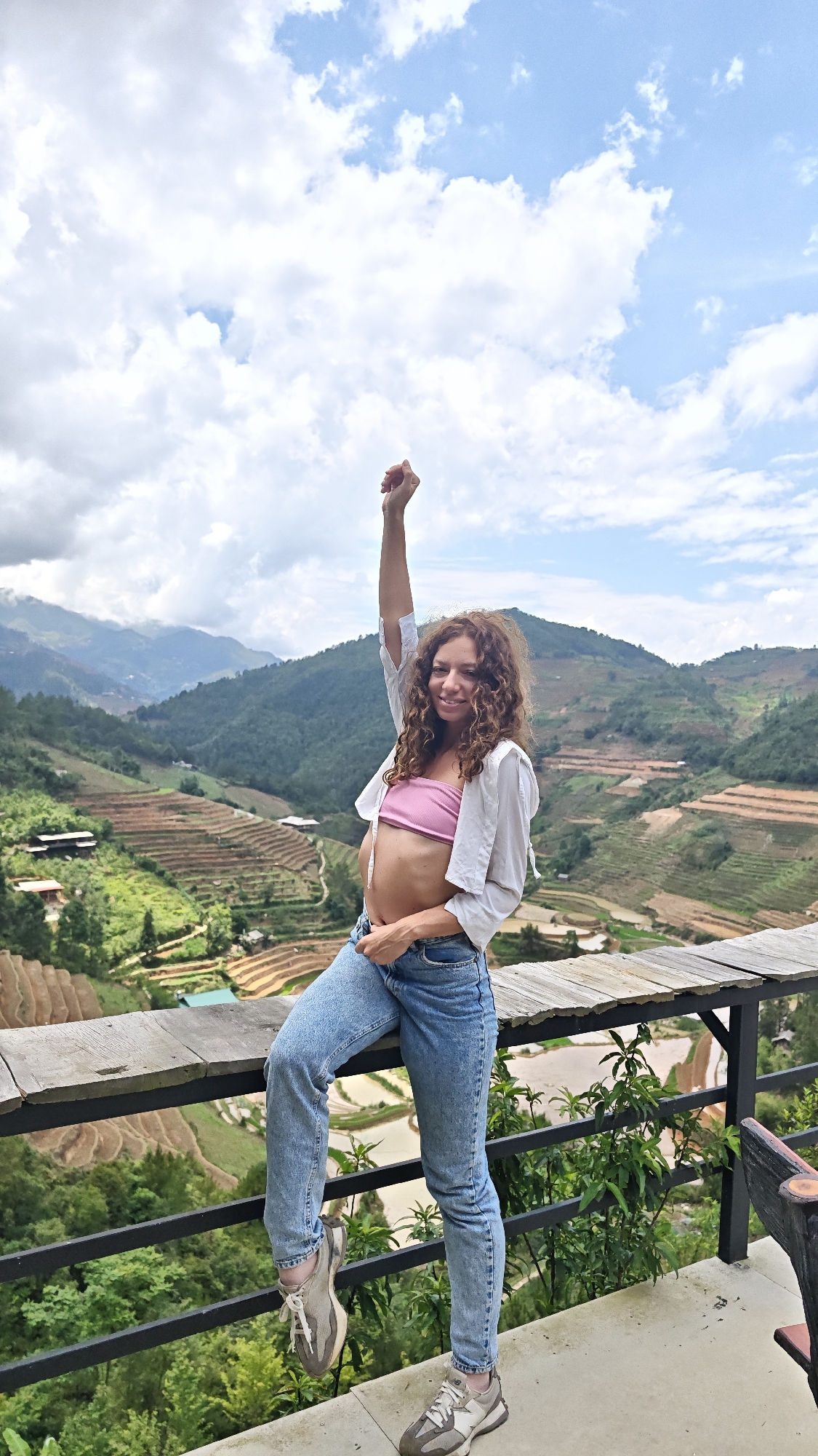 A woman stands on a wooden deck with a metal railing, likely a viewpoint in the city of Ngã Ba Kim, Vietnam. She is wearing a pink tube top, a white button-down shirt, blue jeans, and sneakers. Her right arm is raised in a fist. The background reveals a landscape of terraced rice fields and mountains under a cloudy sky.