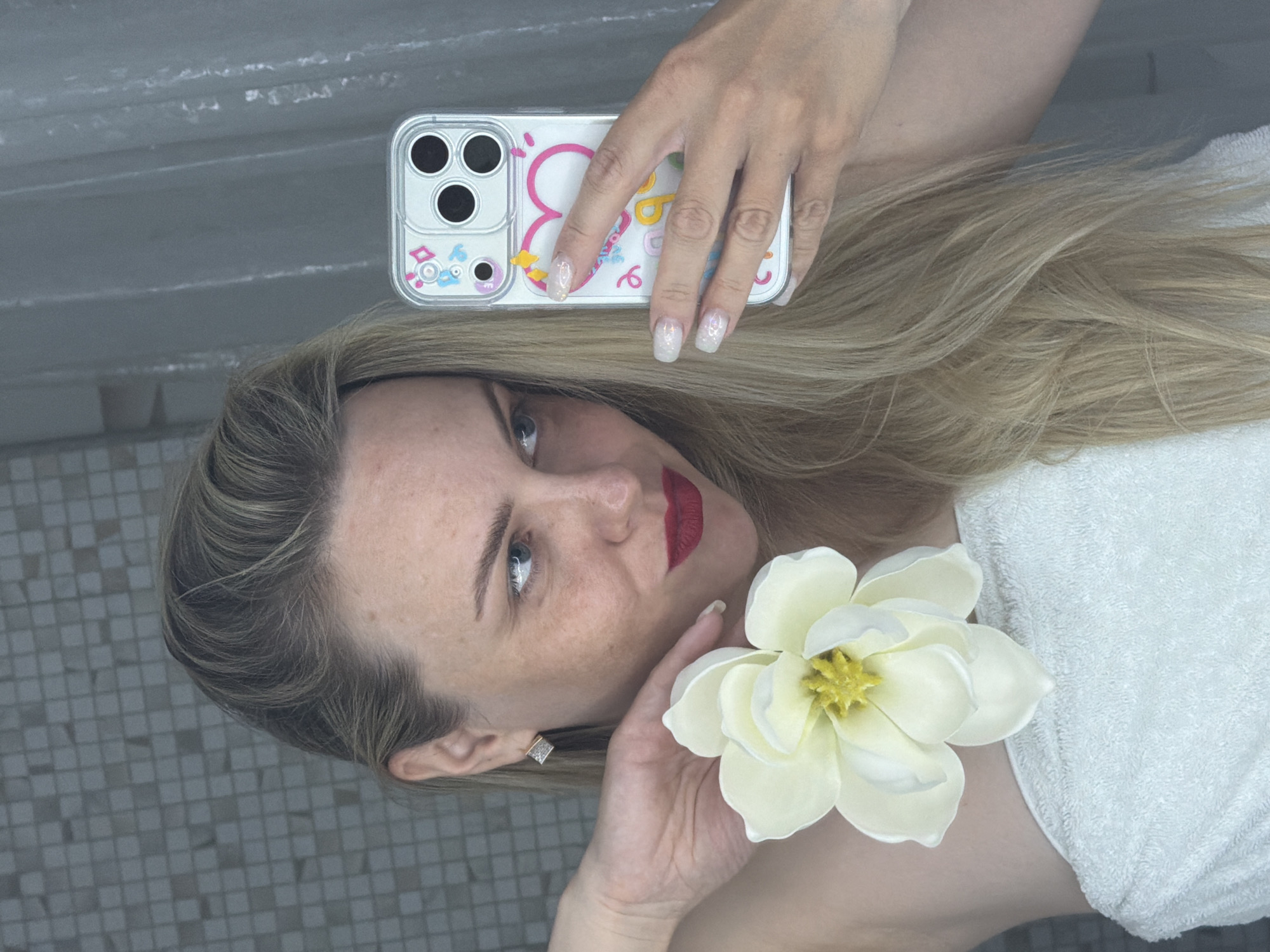 A woman is lying down, holding a white flower and a phone with a decorated case. She is wearing red lipstick and an earring. She appears to be on tiled steps. The location cannot be determined based on the image.
