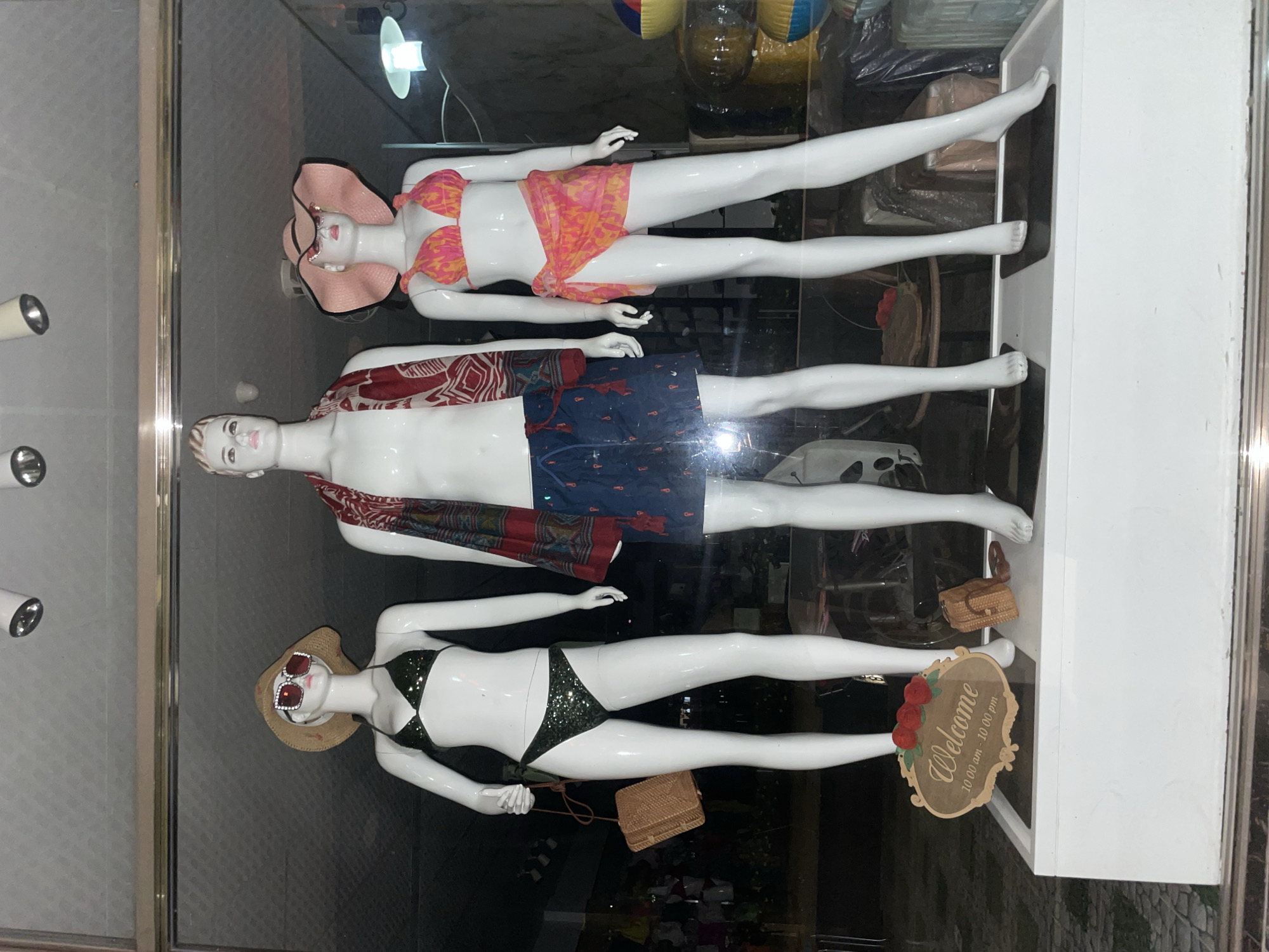 A retail display in a store in Phu Thuy Ward, Vietnam, features three mannequins. The mannequins, all full-body, are positioned horizontally in a glass-fronted display case. The top mannequin wears a pink sun hat and a pink and orange patterned two-piece swimsuit. The middle mannequin, seemingly male, wears a red patterned scarf and blue patterned swim trunks. The bottom mannequin wears a straw hat with sunglasses and a green sequined two-piece swimsuit.
Adjacent to the bottom mannequin is a small straw handbag and a decorative "Welcome" sign, which indicates the store's operating hours are from 10:00 am to 10:00 pm. The display case is enclosed within a white structure. The environment is well-lit, with multiple light sources visible, some reflected in the glass.