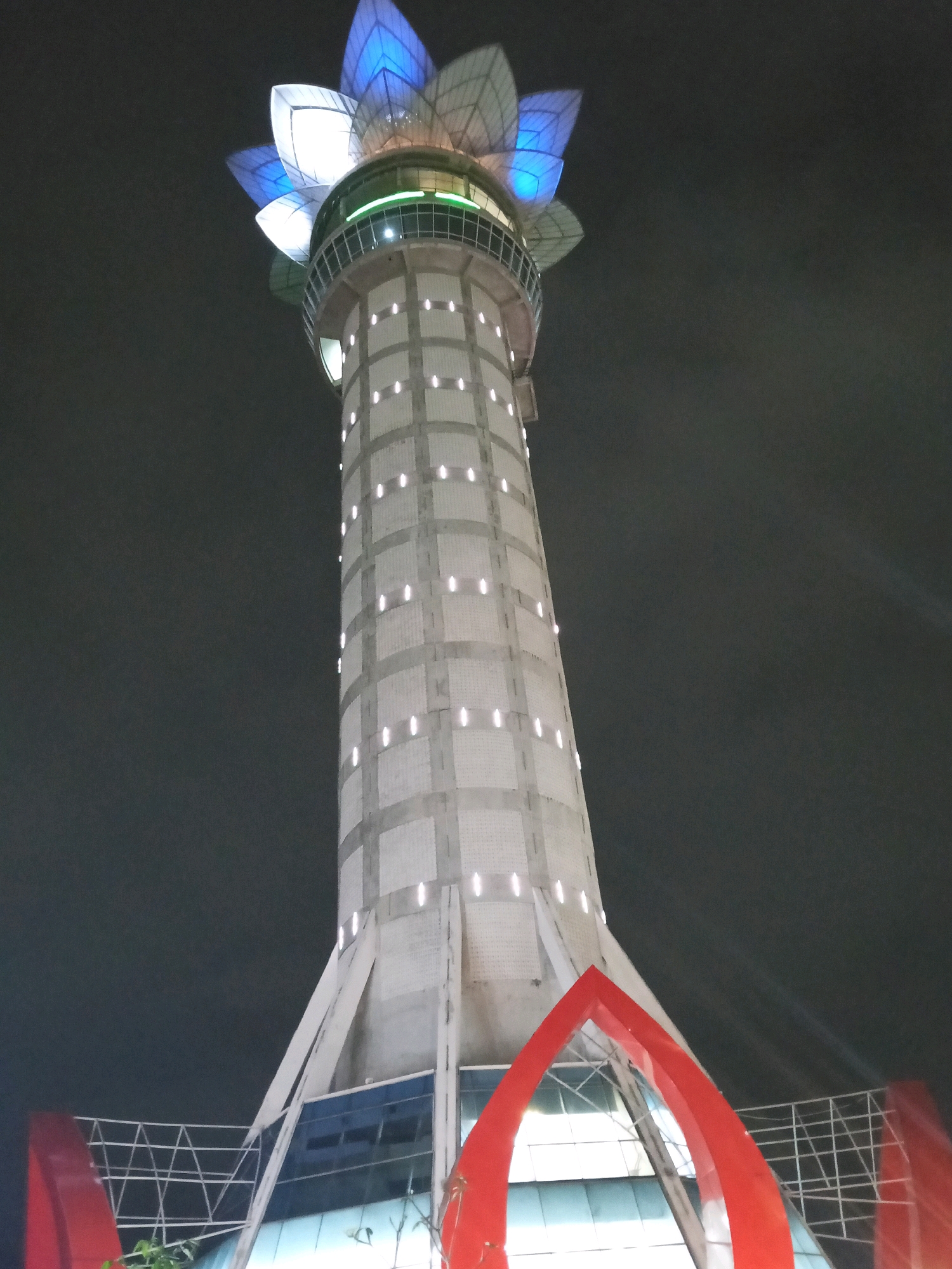 A tall, modern tower with a lotus-like illuminated top stands against a dark night sky. The tower has a cylindrical base, tapering upwards, with a grid pattern of white lights on its facade. The top of the tower features a structure resembling flower petals, illuminated with blue and white lights. At the base of the tower, a modern building with glass windows is visible, and a section of red architectural element curves upwards. This landmark is the Masjid Agung Baitussalam tower in Purwokerto, Indonesia. There are no people visible in the image.