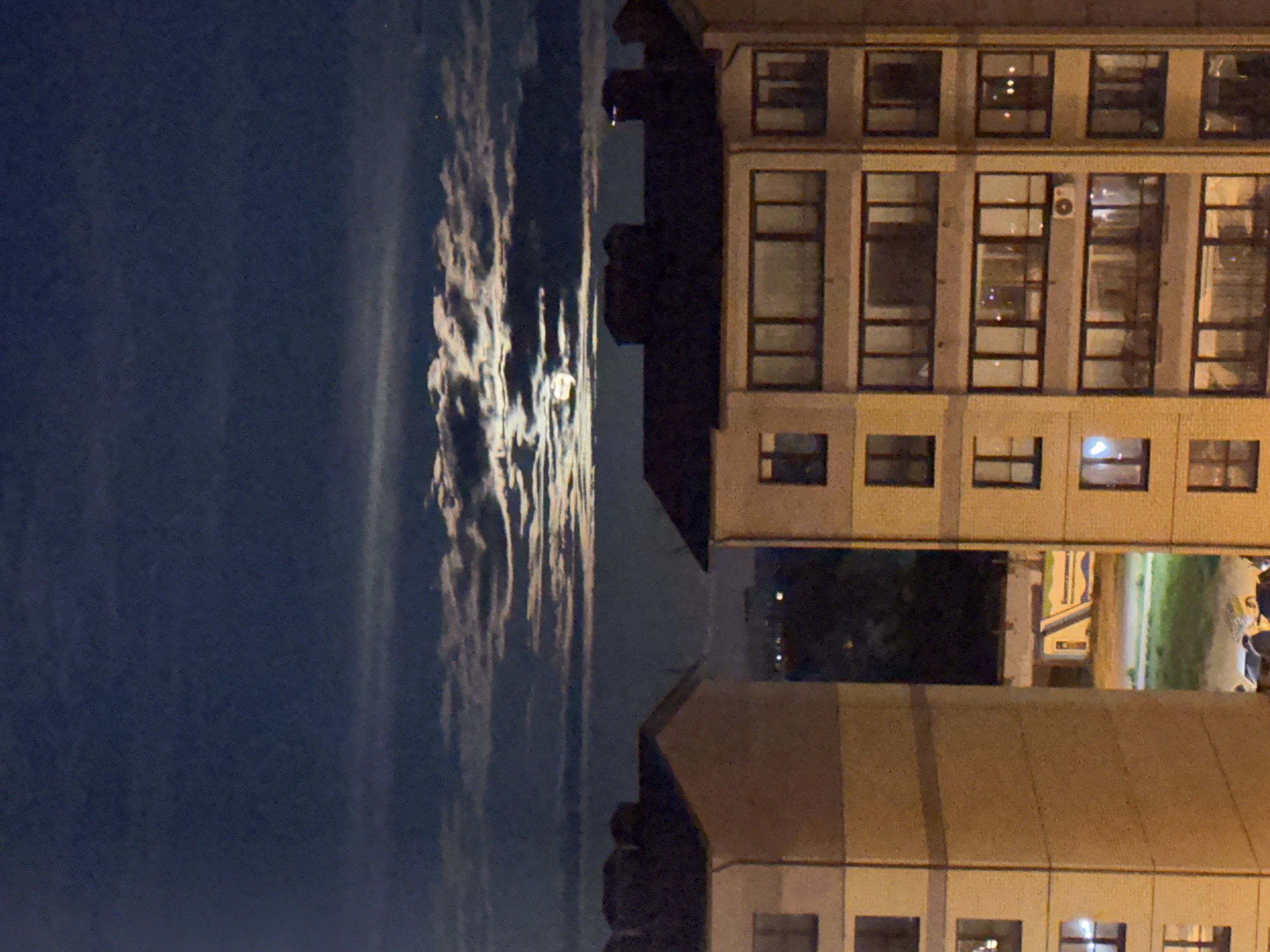 At night in Vladimir, Russia, a building with several lit windows stands to the right. To the left, the moon is partially obscured by clouds. Other buildings are visible in the background.