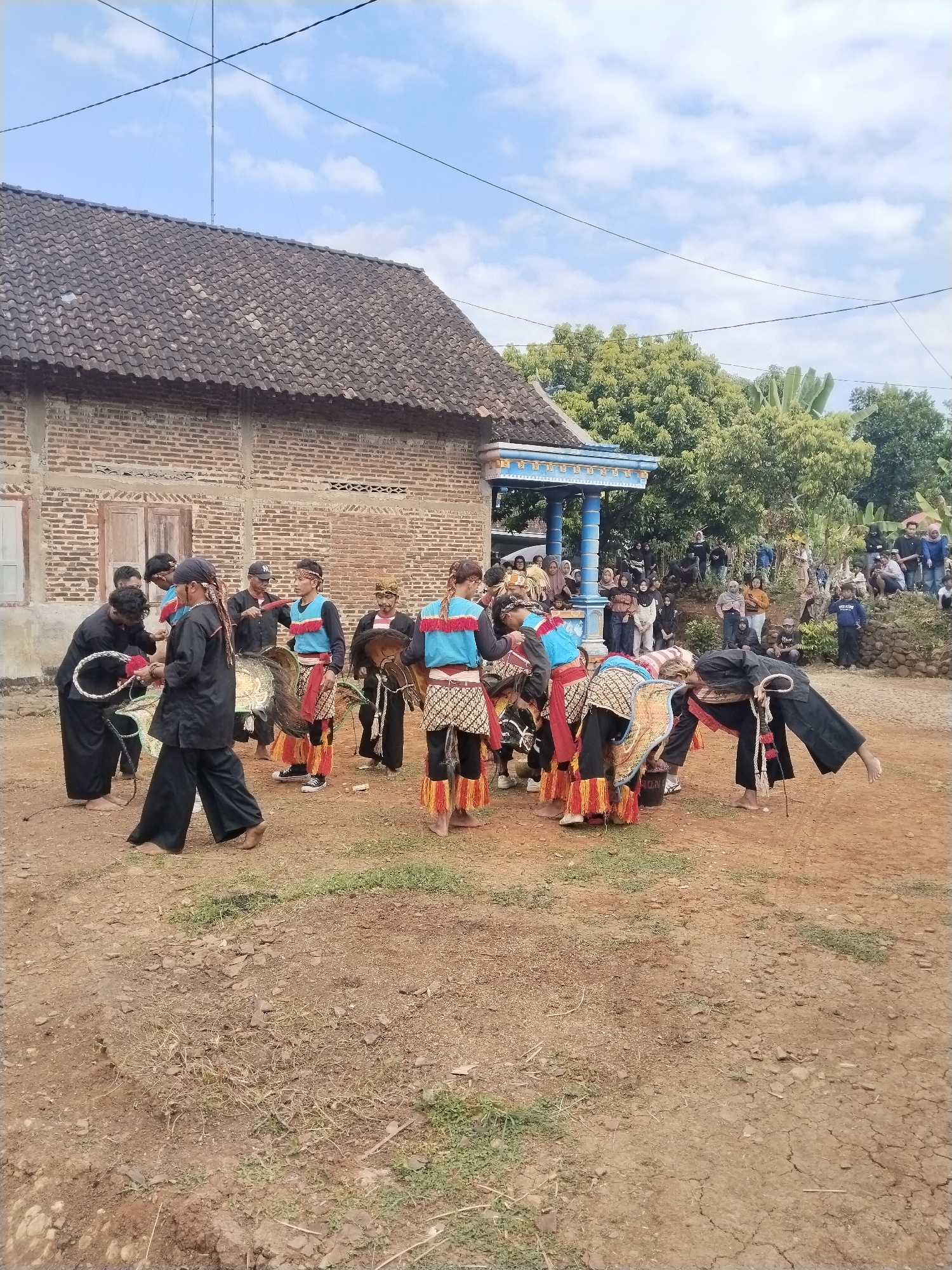 In Gentungan, Indonesia, a group of people are performing a traditional cultural dance. The dancers wear a combination of black, red, blue, and white clothing, and some carry drums or other musical instruments. The performance is taking place on a dirt patch in front of a brick building with a brown tile roof. A crowd of people is gathered behind a decorated gazebo to watch. The sky is partly cloudy.