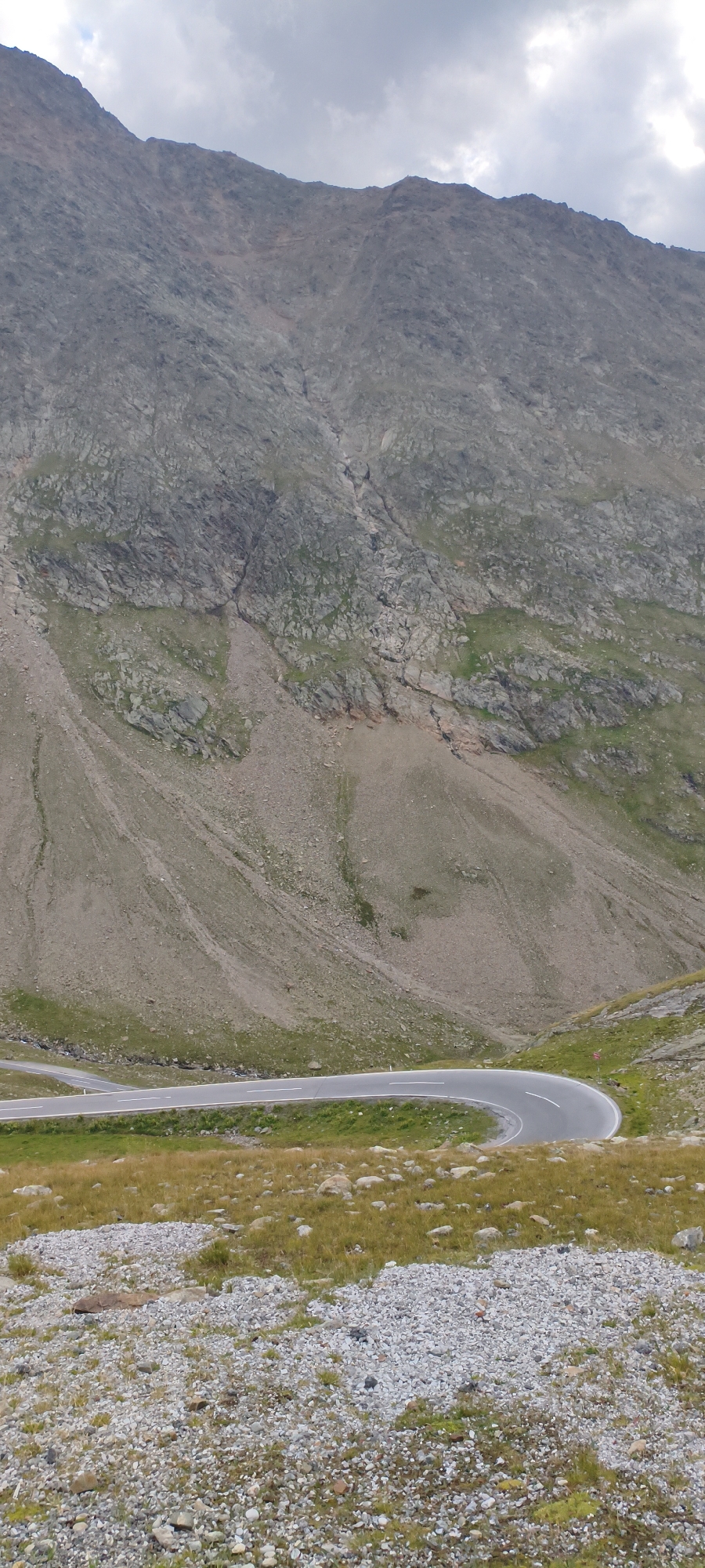 This image captures a winding mountain road in the vicinity of Sölden, Austria, leading up a steep, rocky mountainside under a cloudy sky. The road, paved and marked with white lines, curves sharply through a grassy slope dotted with small rocks and patches of yellowing grass. The mountain itself is a dominant feature, characterized by its rugged, grey and brown rock face, with sparse vegetation clinging to its lower slopes. A visible water or debris channel runs down the mountainside, indicating past erosion. The foreground consists of a rocky, uneven terrain with a mixture of small grey and white pebbles and sparse, dry grass. No people or vehicles are present in the frame.