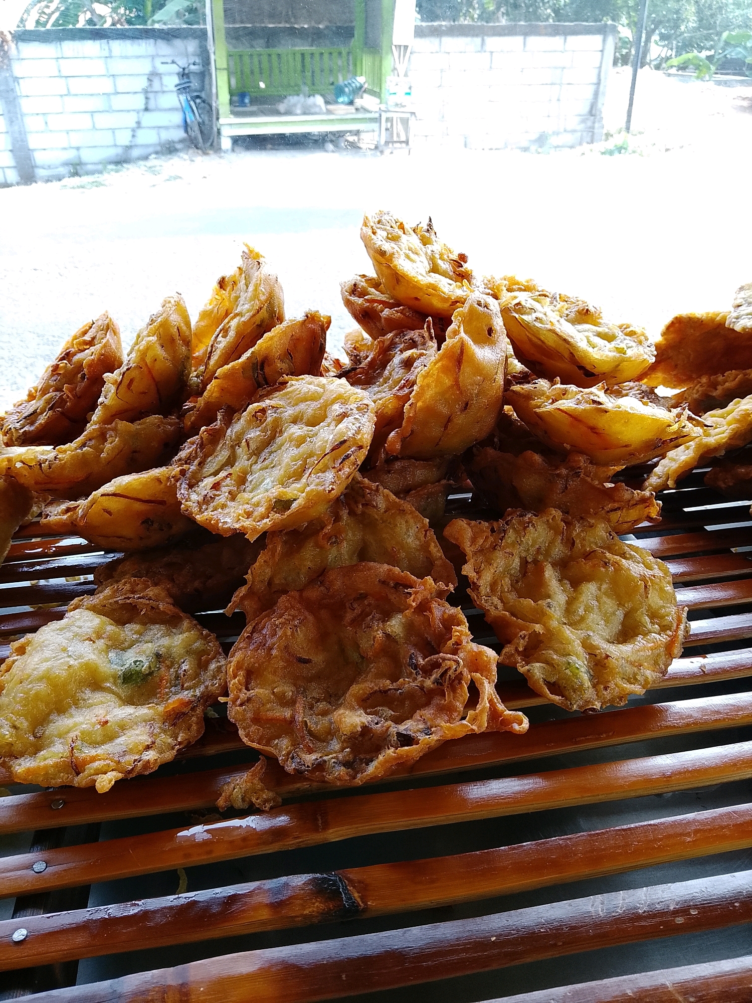 The image shows a close-up of a pile of Indonesian fritters, likely "bakwan," arranged on a bamboo mat. The fritters are golden brown and appear to be made with vegetables, possibly onions and carrots, mixed with a batter and deep-fried. They are stacked in a casual, inviting manner. In the background, a blurred view of an outdoor setting is visible. This appears to be a stall or a roadside eatery in Banaran, Indonesia, as indicated by the general appearance of the structures and vegetation. The overall scene suggests a simple, local food vendor.
