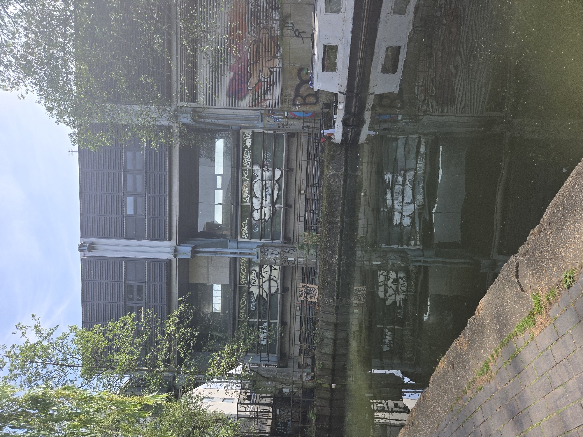 The image shows a canal in an urban setting, likely in London, United Kingdom, given the architectural style. A modern building with a louvered facade stands adjacent to the canal. Graffiti is visible on the building's facade and surrounding structures. The building is reflected in the canal's water, which also reflects other elements of the scene. A narrowboat is moored on the canal. Trees with green foliage are partially obscuring the building. The towpath of the canal is lined with brick and concrete.