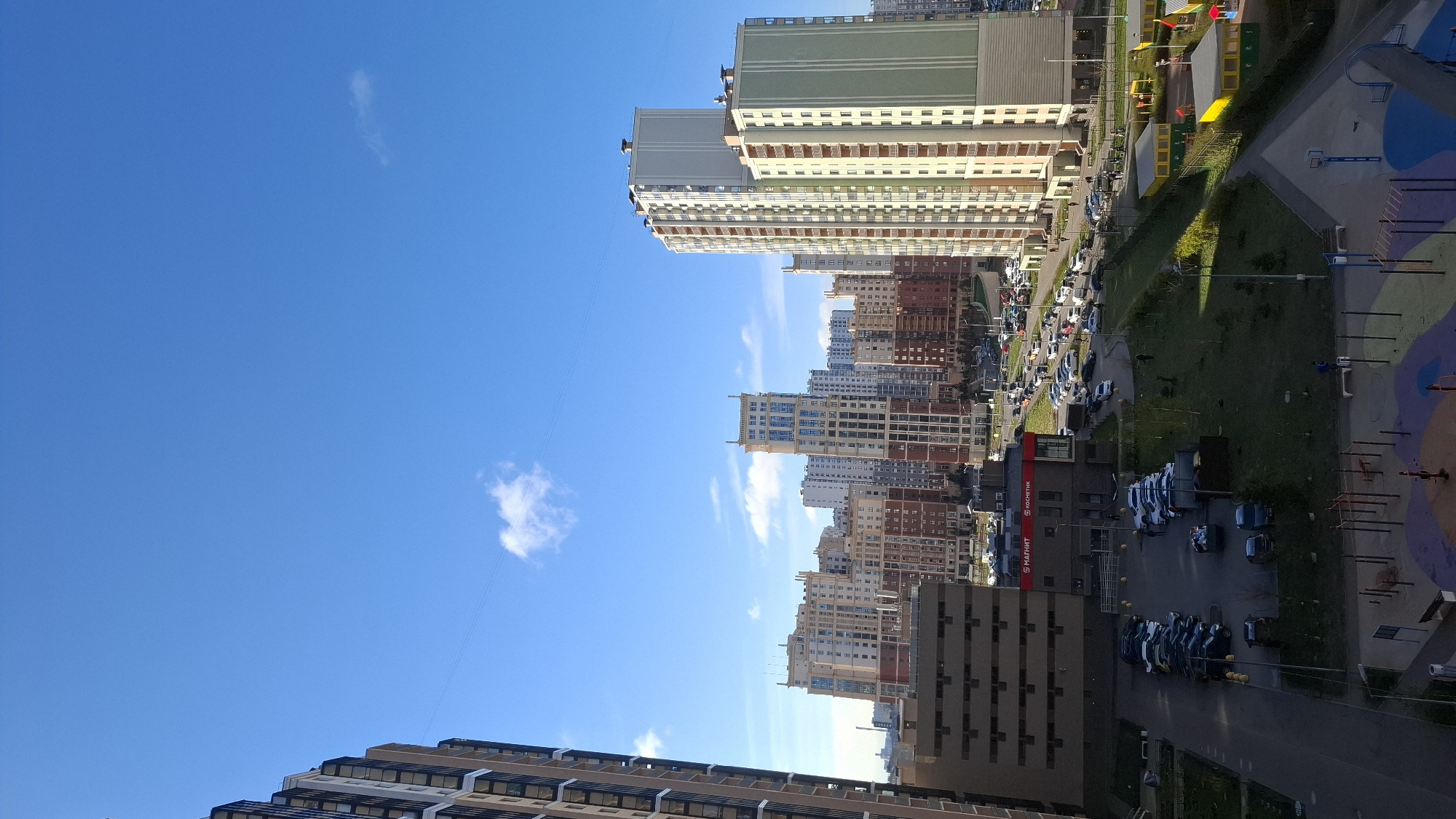 The image captures an expansive, sunlit urban landscape from a high vantage point, looking down upon a dense residential district in what appears to be Saint Petersburg, Russia. The perspective is tilted, showing tall modern buildings stretching towards a bright blue sky with a few wispy white clouds.
The scene is dominated by numerous high-rise residential apartment buildings, varying in height, architectural style, and façade colors, including shades of beige, brown, grey, green, and reddish-brown. Many buildings feature multiple balconies and windows. In the foreground and middle ground, building clusters create a sense of a tightly packed urban environment.
Below the buildings, the ground level reveals streets and pathways bustling with everyday activity. Numerous cars are parked along roadsides and in dedicated lots, suggesting a residential area with a high density of vehicles. A few cars are visible on the roads, indicating ongoing traffic. Patches of green lawn and scattered trees provide natural elements amidst the concrete and asphalt.
To the right, a colorful playground or recreational area with distinct shapes and possibly equipment is visible, hinting at family-friendly amenities. Power lines stretch across the clear sky, connecting infrastructure. Shadows cast by the buildings suggest it is either morning or early afternoon on a clear, sunny day.
Visible text on a red building in the mid-ground includes "5 МАГНИТ" (likely a "Magnit" supermarket, a common chain in Russia) and "КОСМЕТИК" (Cosmetics), confirming commercial activity within this residential setting. The overall impression is that of a modern, well-developed urban residential area under clear skies.