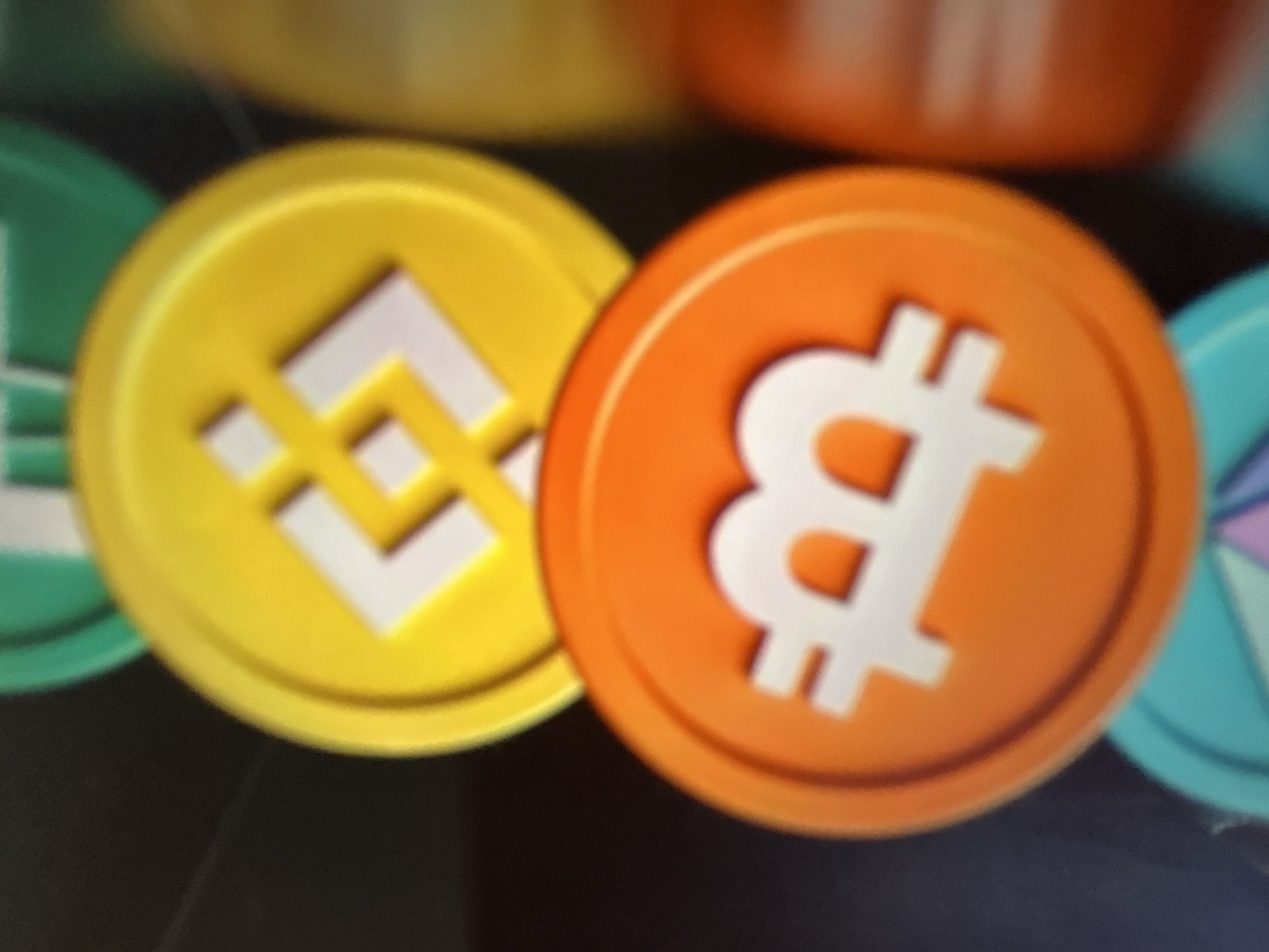 This media file is a close-up, slightly blurry photograph of several cryptocurrency coins. The primary subjects are a yellow coin with a white logo that appears to be the Binance logo, and an orange coin with a white Bitcoin logo. These coins are arranged next to each other, with the yellow coin partially overlapping the orange coin. There are also hints of other coins in the background, one appearing to be green and another teal with a geometric design. The coins are rendered in a 3D style, with raised logos and beveled edges, suggesting they might be digital assets or tokens. The background is dark and out of focus, providing no specific setting details. The lighting suggests an indoor environment, possibly a desk or a display area for digital currency-related items. There is no discernible text in the image other than the cryptocurrency logos themselves. The image does not depict people or a specific event. The overall impression is a visual representation of popular cryptocurrencies.