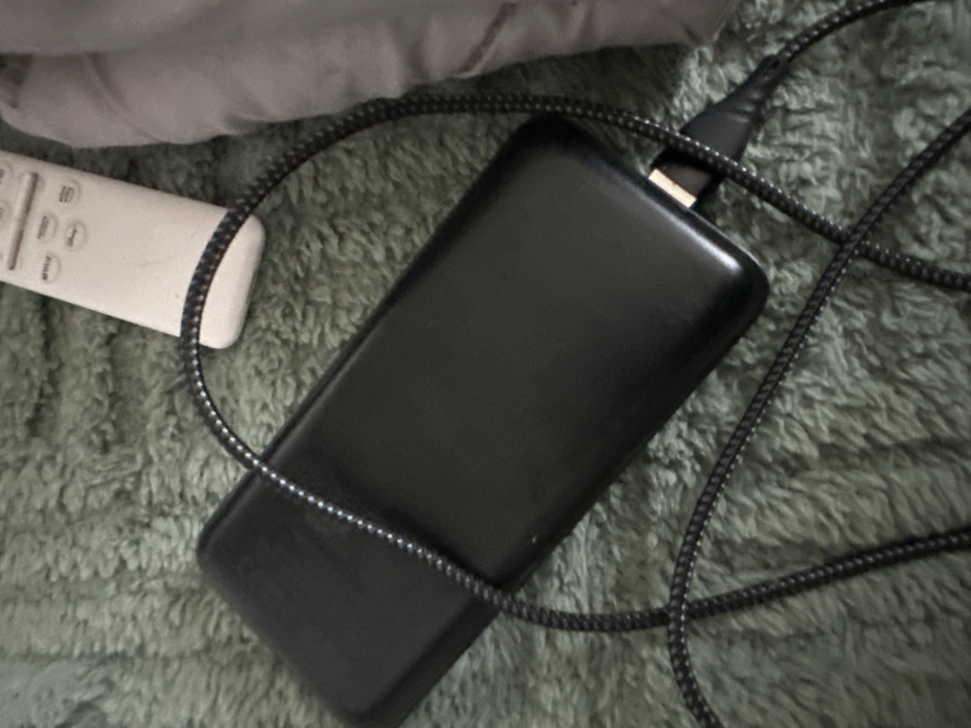 The image shows a black rectangular power bank laying on a fuzzy green blanket or surface. A black braided charging cable is connected to the power bank. A white remote control is partially visible to the left of the power bank, near the top of the image. A portion of a gray cushion or piece of clothing is visible above the remote control. The photo provides no information to suggest the location is Slavyani, Bulgaria.