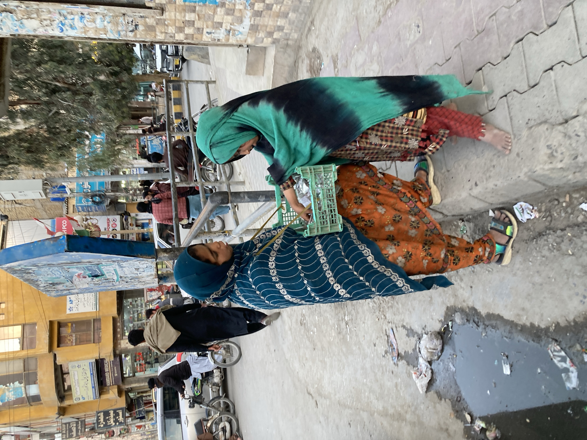 Two women are sitting on the side of a street in Quetta City Tehsil, Pakistan. One woman wears a green and black shawl and a red and brown patterned garment. The other woman wears a blue patterned garment and an orange patterned garment, and holds a green plastic crate. The women are barefoot or wear sandals. The street is cluttered with litter, and there is a puddle of water near the women. In the background are buildings, signs, and people on bicycles and motorcycles.