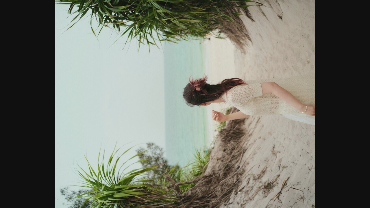 A woman, viewed from behind, stands on a sandy path leading to a beach. She has dark hair pulled up in a bun with a clip. She is wearing a cream-colored, short-sleeved, crocheted dress over a white underskirt and a pearl bracelet. The path is flanked by sandy dunes covered in dry vegetation and palm-like plants. The ocean is visible in the background under a hazy sky. The scene appears to be located at a beach, with the specific city and country unknown.