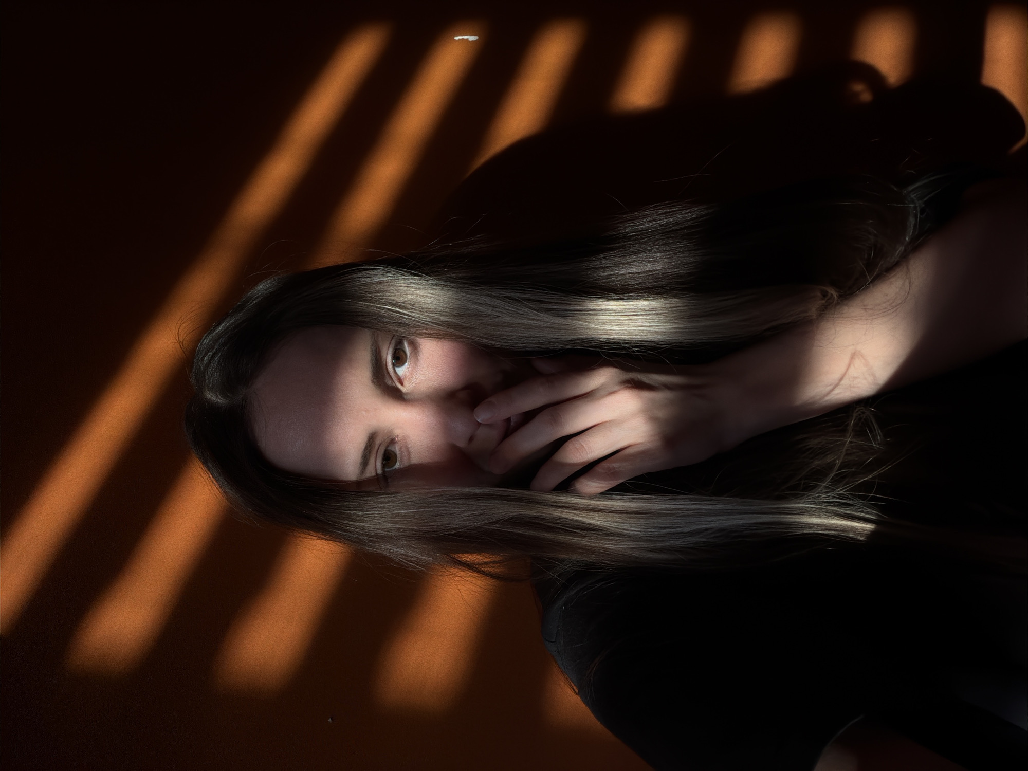 A female individual is depicted in Khimki, Russia, seated indoors against a plain, warm orange-brown wall. She has long, straight, multi-toned hair, predominantly dark brown with lighter blonde or grey streaks, particularly visible on the left side of her head. Her right hand is raised, with fingers covering her mouth and the lower portion of her nose. Her right eye, brown in color, is partially visible and directed towards the viewer. The left side of her face is obscured by hair and shadow. She is wearing a dark, short-sleeved garment, possibly a t-shirt. The scene is lit by strong, directional light, casting distinct horizontal bands of illumination and shadow across the wall and the subject, indicative of light filtered through window blinds or slats. Significant portions of the subject, including her left side and much of her clothing, remain in deep shadow.