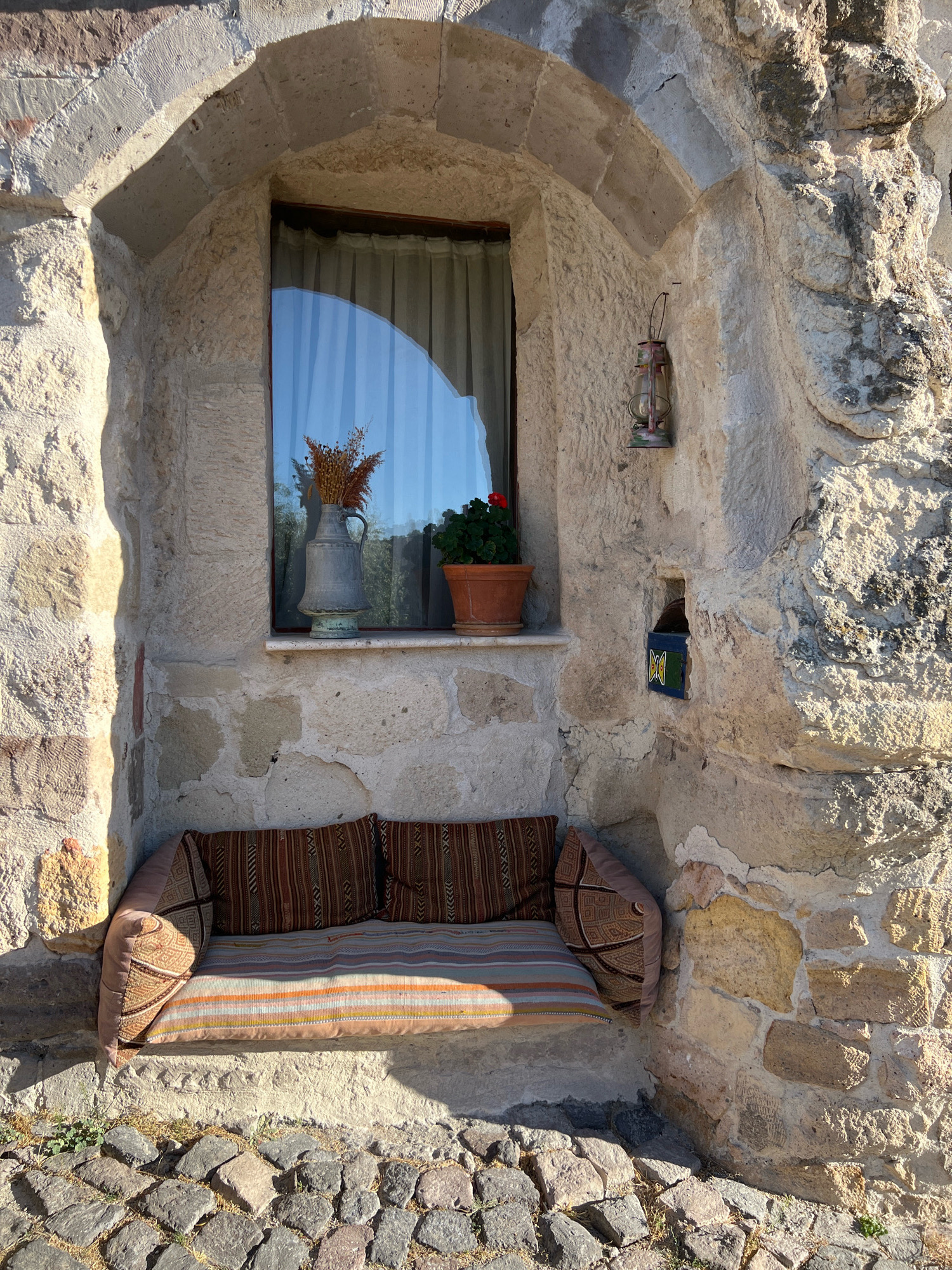 The image depicts a cozy niche built into a stone wall in Göreme, Turkey. A window with sheer curtains is centrally located, reflecting a clear blue sky. On the windowsill, a vintage metal pitcher holds dried flowers, and a potted red flower adds a splash of color. Below the window, a cushioned bench with patterned textiles invites relaxation. To the right of the window, an old oil lantern hangs from the wall. Further to the right, a small, cubby-like recess in the stone wall contains a colorful tile with a geometric pattern. The ground in front of the niche is paved with cobblestones. The stone architecture is rough-hewn and rustic, typical of the region. The lighting suggests it is daytime, with sunlight casting shadows.