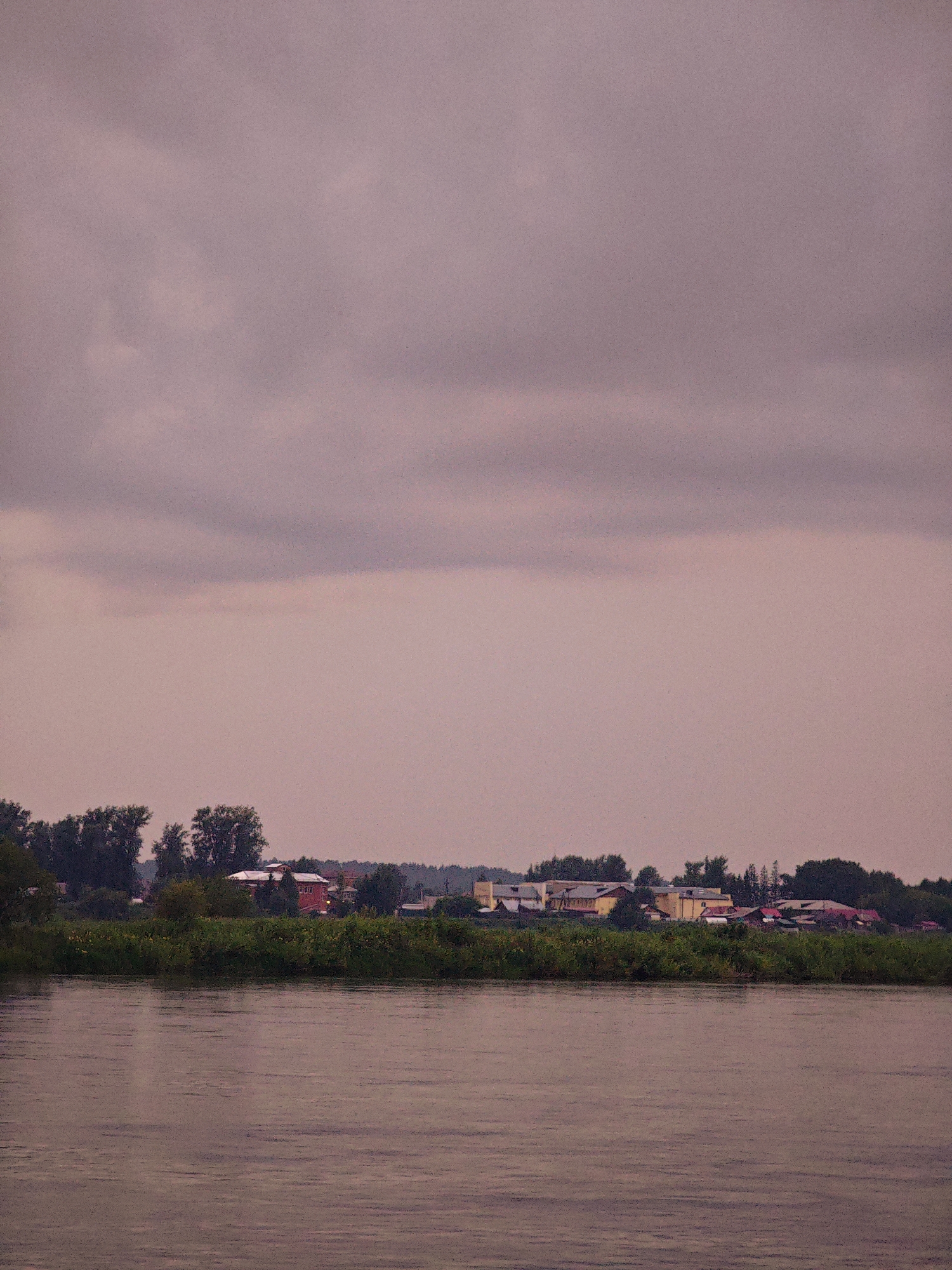 The image depicts a tranquil river scene with a distant village on the opposite bank. The sky is overcast with muted, purplish clouds. The river in the foreground is calm, with subtle ripples reflecting the soft light. A dense line of green foliage, likely trees and bushes, separates the river from the village. The village itself is comprised of several buildings, including a prominent red structure and lighter-colored buildings with red roofs. The architecture suggests a rural or semi-rural setting. The overall mood of the image is serene and somewhat somber due to the cloudy sky. The location is the city of Kazachinskoye, Russia.