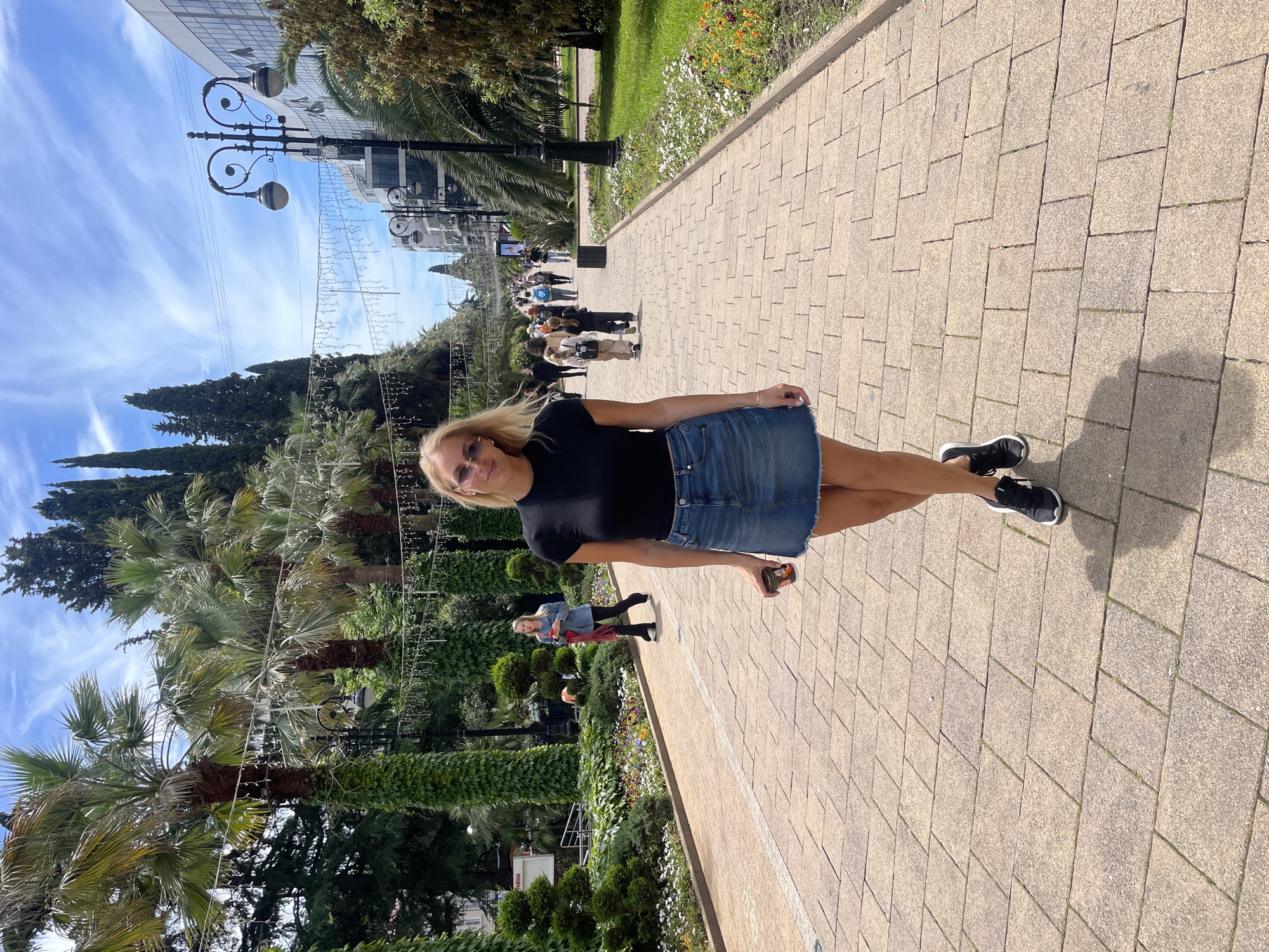 A photograph taken outdoors shows a woman in a denim skirt and black top standing on a brick walkway in Sochi, Russia. She is wearing sunglasses and black sneakers. To her left are palm trees and other greenery; to her right is a path lined with flowers. In the background, people walk along the path, and a building and streetlights are visible. The sky is blue with scattered clouds.
