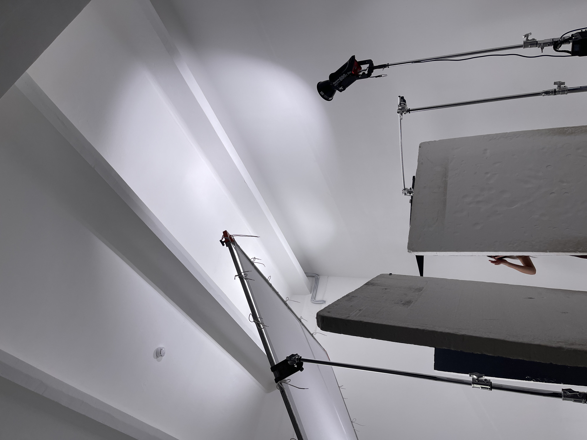 This media file captures an indoor photography or video studio setup, characterized by white walls and a sloped white ceiling with visible structural beams. The scene shows various pieces of professional lighting and light-shaping equipment, indicating a shoot is either underway or being prepared.
In the upper-right section, a black Aputure 600d Pro light fixture is mounted on a boom arm, directed towards the ceiling. Below this, two large, rectangular panels, possibly V-flats or bounce boards, are suspended horizontally. The panel closer to the foreground is dark gray and textured, while the one behind it is lighter in tone. A pair of human hands and forearms are visible between these two suspended panels, suggesting a person is present and actively involved in the setup or interaction with a subject.
On the left side of the frame, a large light diffuser or reflector screen is positioned at an angle, supported by a stand and boom arm. The bright white ceiling also features a small, circular smoke detector. The metallic support arms, clamps, and light stands are prominent throughout the image. The controlled environment and specialized equipment suggest a professional creative project in progress within a studio setting, potentially located in Moscow, Russia.