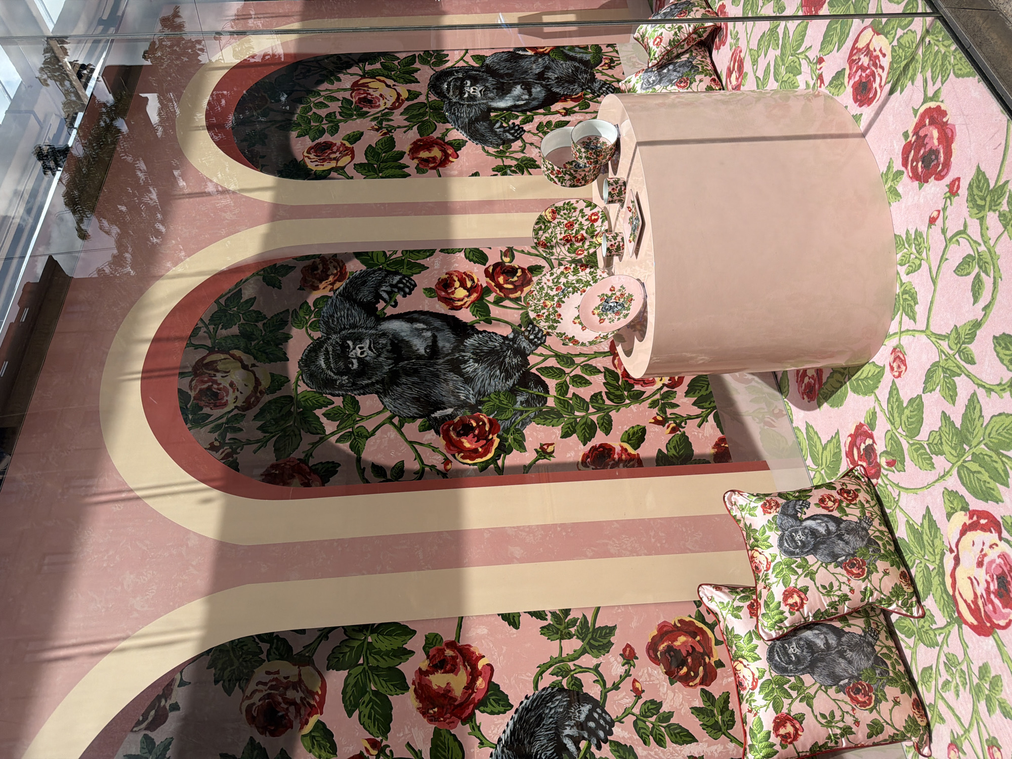 This eye-level photograph shows a Gucci display inside of a storefront window in Milan, Italy. The store design features a pink palette with floral and gorilla-themed decor. Arched, striped patterns run across the window's background, with floral and gorilla prints filling the spaces in between. A pink, cylindrical table sits centrally in the display, with a tea set with matching floral and gorilla prints on top. The set includes cups, saucers, and small plates. Two throw pillows, matching the prints on the floor, are in the bottom right corner. Outside, the reflections of trees and building facades indicate a street scene with natural light. The glass storefront slightly distorts and reflects the outside environment, offering a glimpse of the city's architecture and foliage.
