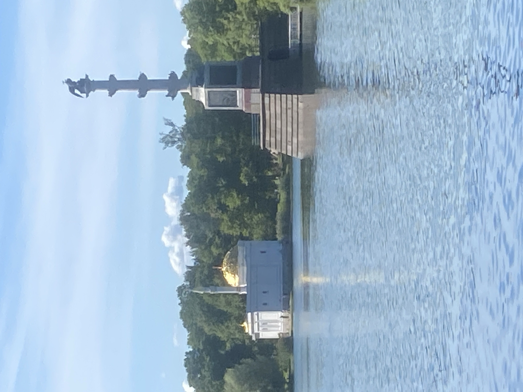 The image shows a serene lake with a white building topped with a golden dome on the left, and a tall, multi-tiered monument topped with a statue of a bird on the right. Both structures are situated near the shore, surrounded by lush green trees. The sky above is clear blue with a few wispy clouds. The water of the lake is calm, reflecting the sky and the surrounding scenery. This scene is likely from the Catherine Park in Pushkin, Russia, a renowned landmark famous for its picturesque landscapes and architectural marvels.