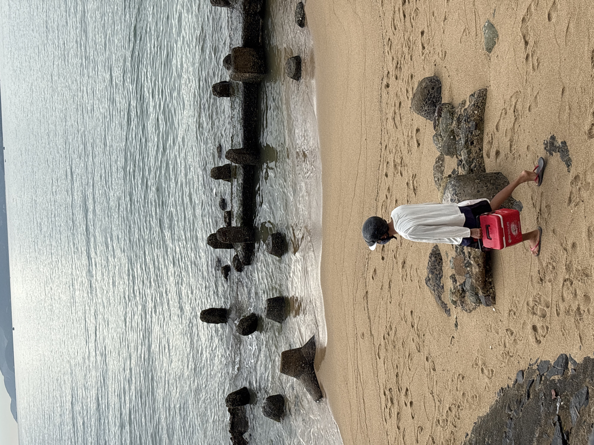 An individual is walking on a sandy beach in Nha Trang, Vietnam, carrying a red cooler bag. The individual is wearing a white long-sleeved shirt, dark shorts, a helmet, and flip-flops. The beach is bordered by the ocean, which has a breakwater made of concrete structures and rocks. In the distance, mountains are visible on the horizon. The sand is imprinted with footprints. The overall lighting suggests an overcast day.