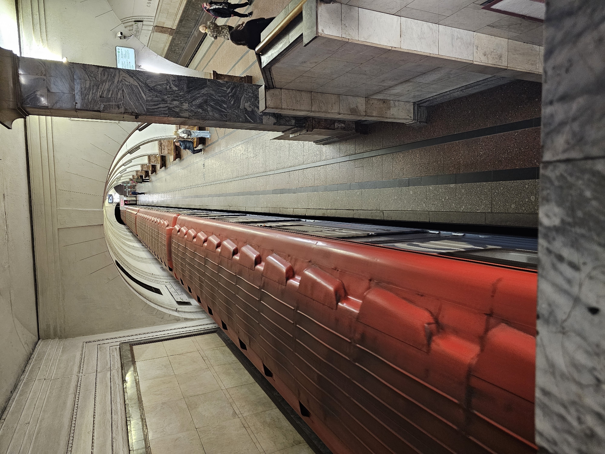 The image depicts a red train in a Moscow Metro station, Russia. The station features marble walls and columns. Passengers are seen waiting on the platform. The camera angle is looking down on the train.