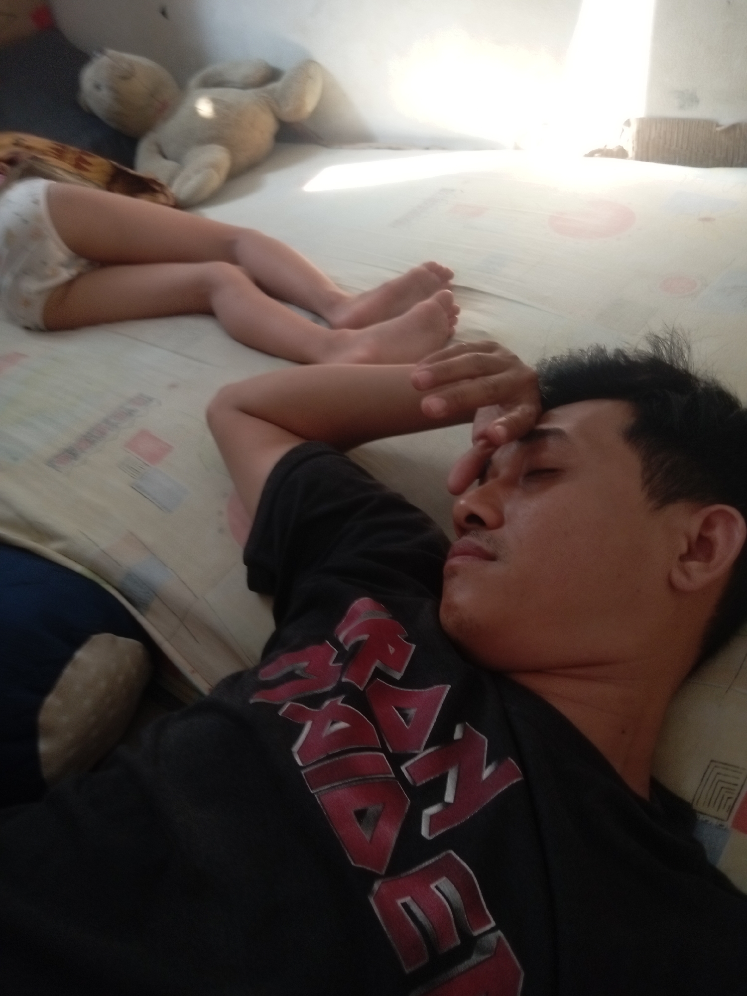 A man is lying down, seemingly asleep, with his hand on his forehead, on a white mattress that appears to be in a bedroom. Beside him, a baby's legs are visible, suggesting the man might be a father resting while his child is nearby. In the background, a stuffed toy can be seen, adding a domestic element to the scene. The room is dimly lit, with sunlight filtering in from the right side, creating a warm and soft ambiance. The overall impression is one of peace and relaxation.