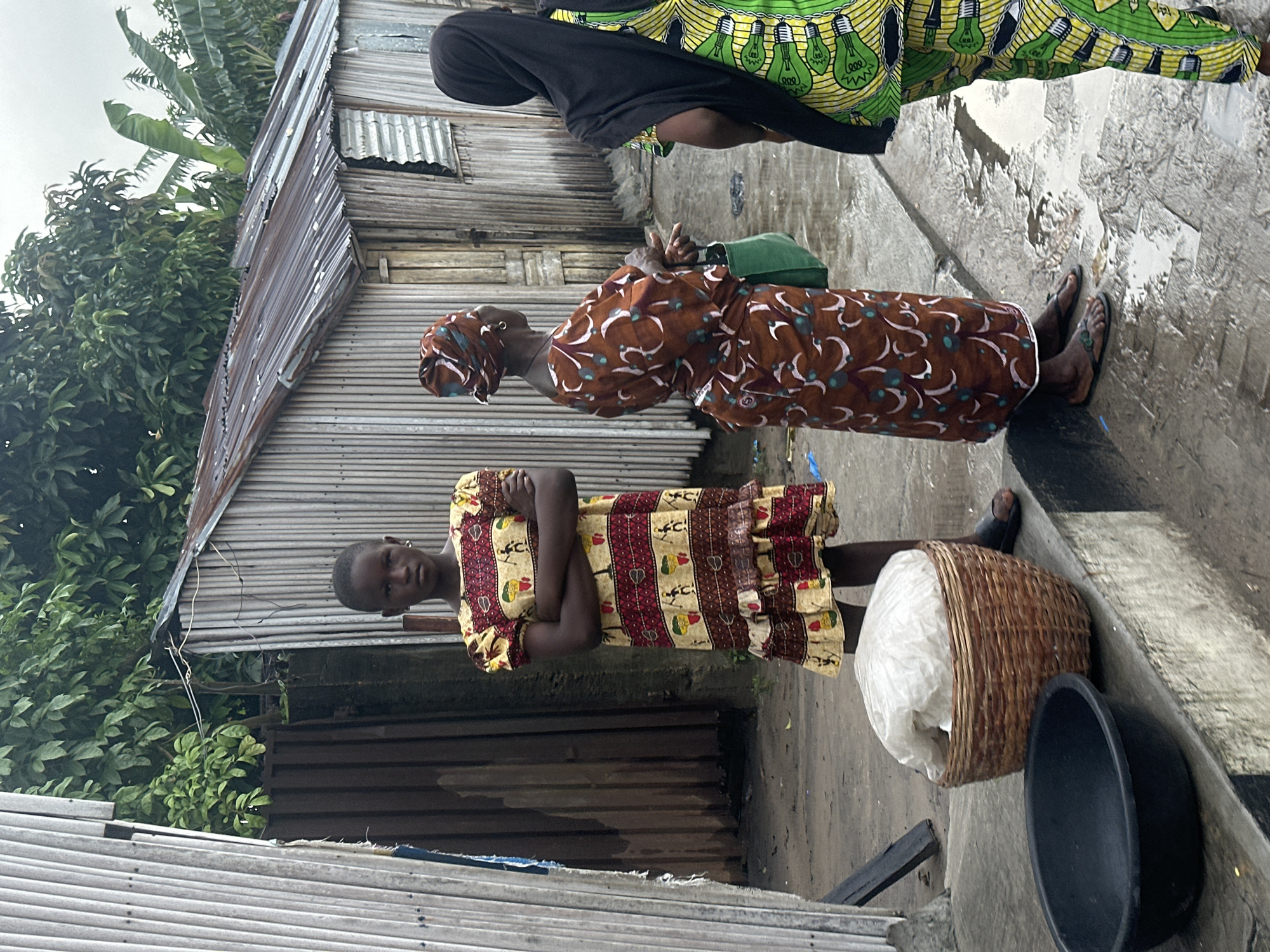 In Badagry, Nigeria, a young girl stands with her arms crossed in front of a weathered corrugated metal structure, her patterned dress featuring earthy tones and depictions of figures. To her right, an older woman, also adorned in a patterned garment and headscarf, faces away from the camera, holding a green bag. Further to the right, another individual, veiled in black, wears a vibrant green and yellow outfit with a lightbulb motif. The ground is wet, suggesting recent rain, with puddles reflecting the overcast sky. A woven basket containing a white bundle and a dark basin are placed on a concrete ledge in the foreground. The setting appears to be a modest, possibly informal, area with elements of urban decay and lush vegetation in the background.