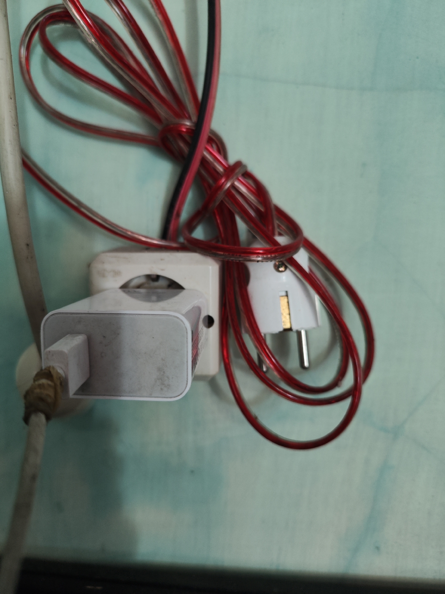 A white electrical adapter is plugged into a white wall socket. A white cable is connected to the adapter and extends out of frame to the left, secured by a tan colored tie. A coiled red and black wire bundle is also present, with a white plug attached to it inserted into another socket, or possibly the same one if it's a dual socket. The wall behind the electrical components is a light blue-green color and appears to be textured or stained. This image was taken in Bulurejo, Indonesia.
