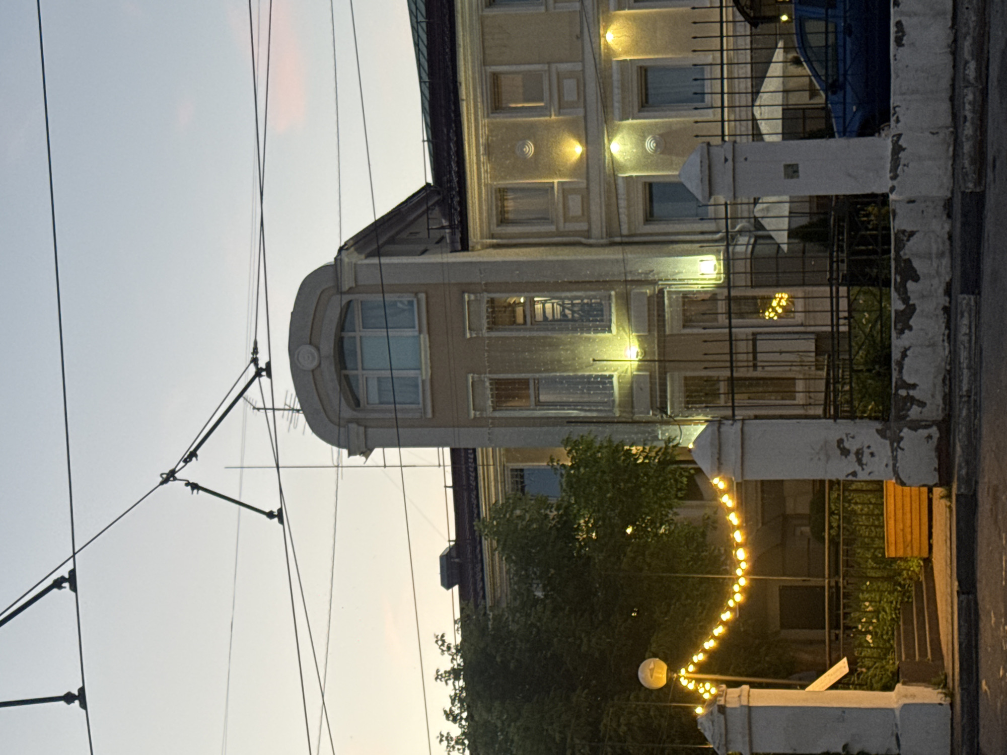 The image depicts a building at twilight in Nizhny Novgorod, Russia, with overhead tram wires crossing the sky. The building, painted in muted yellow tones, features a prominent arched section with a multi-paned window at the top and smaller rectangular windows below. Decorative lighting illuminates the facade, casting a warm glow on the textured walls. A dark metal fence encloses the property, behind which are white umbrellas and a blue car. In the foreground, a string of yellow lights hangs between two white, weathered pillars, spanning over a set of stairs leading to the entrance. A wooden planter box sits to the right of the stairs. The sky is a pale grey with hints of pink, suggesting the end of the day.
