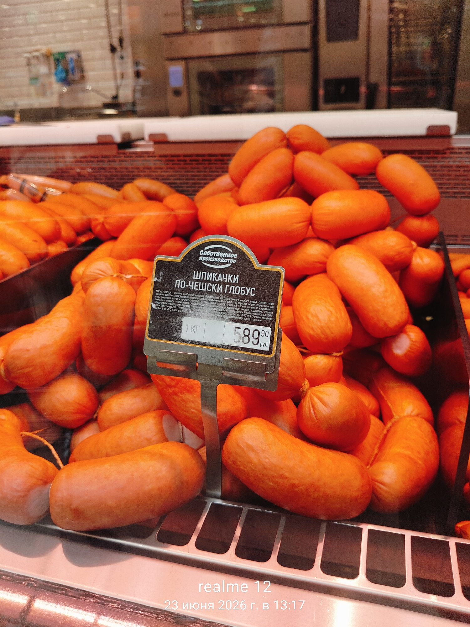This is a close-up, eye-level shot of a refrigerated display case filled with numerous short, plump, bright orange sausages. A dark-colored label with white and yellow text is prominently displayed among the sausages. The label contains Russian text and reads "Собственное производство" (Own production) at the top, followed by "ШПИКАЧКИ ПО-ЧЕШСКИ ГЛОБУС" (Lard sticks Czech-style GLOBUS). Below this, there is a barcode and pricing information including "1 кг" (1 kg) and "589.90 руб." (589.90 rubles). The sausages are tied in pairs with thin string. The background is blurred, showing kitchen equipment and tiled walls, suggesting a food service environment. The bottom right corner of the image displays a timestamp: "realme 12 23 июня 2026 г. в 13:17". This indicates the photo was taken on June 23, 2026, at 13:17, on a device identified as "realme 12".