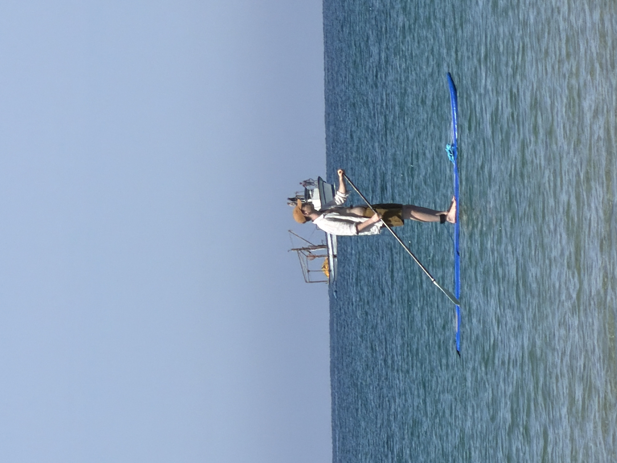 A person in a hat, light-colored long-sleeve shirt, and shorts is stand-up paddleboarding on the Mediterranean Sea, possibly off the coast of Tel-Aviv, Israel. They are holding a paddle and facing the water, with a boat visible in the background. The sky is clear and blue.