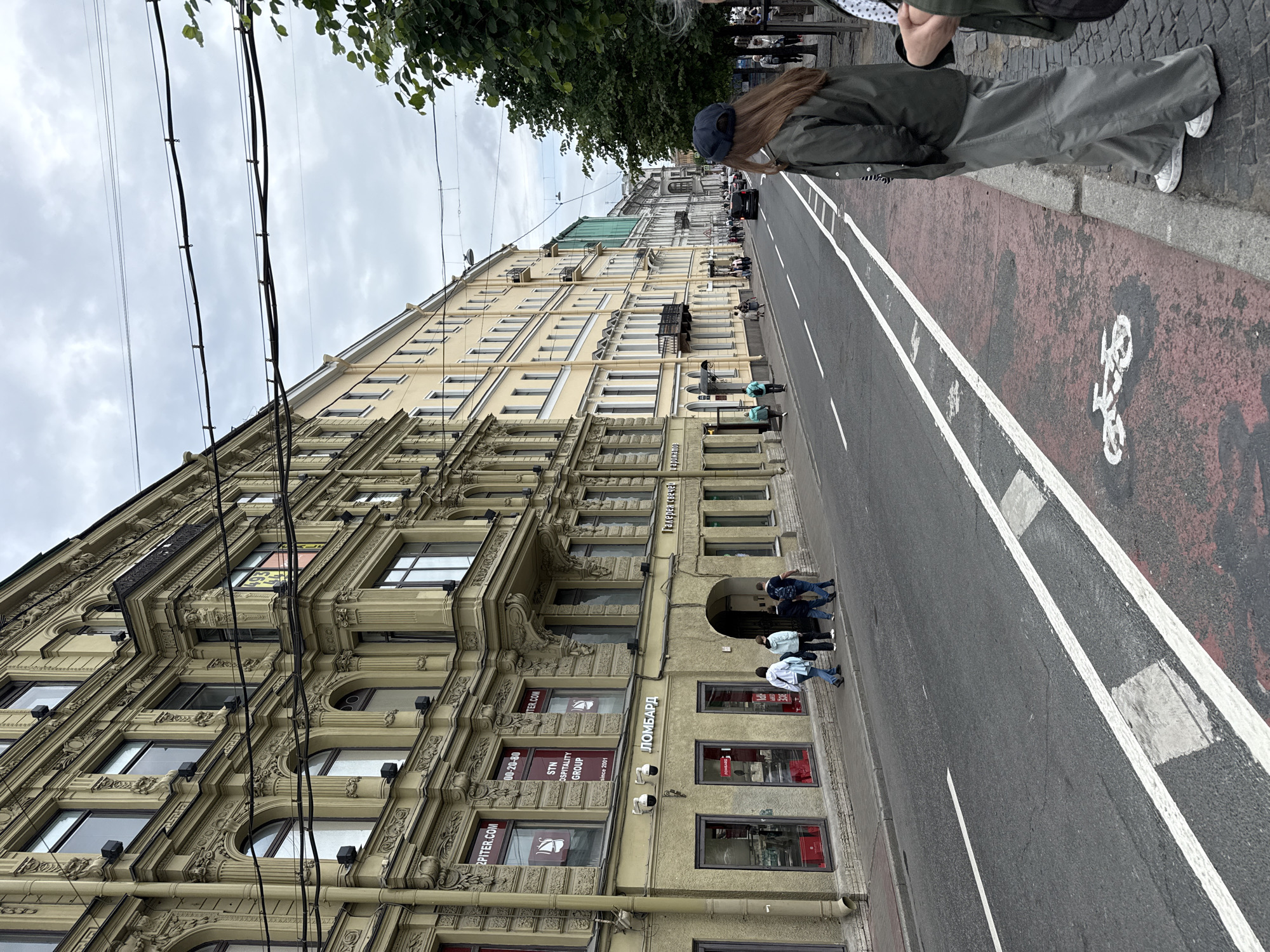 The image depicts a street scene in Saint Petersburg, Russia, featuring historic buildings lining a wide asphalt road. The buildings are characterized by classical architecture, with ornate facades, arched windows, and balconies. The street is lined with trees on the right side, and overhead wires crisscross the sky. Several pedestrians are visible walking along the sidewalk and crossing the street. One person, a young woman wearing a dark cap and a green jacket, stands on the right side of the frame, facing away from the camera and towards the street. The road has white lane markings and a bicycle symbol painted in red on the right side. The sky is overcast, contributing to a muted color palette. The overall impression is of a busy urban environment with a blend of old-world charm and modern city life.