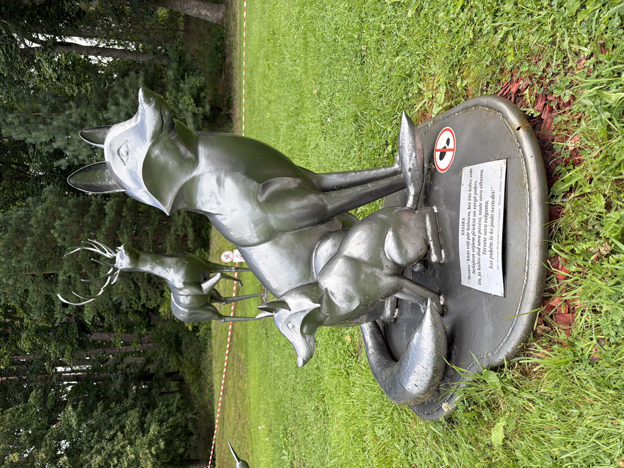 The image shows a sculpture of two foxes, one larger and one smaller, in a grassy area with trees in the background. The foxes are made of a metallic material and are positioned on a circular base. A sign with a red circle and a diagonal line through a footprint is visible on the base. There is a plaque with text on the base, below the foxes. In the background, a sculpture of a deer is visible, as well as red and white tape. This scene is located in the Tērvete Nature Park in Tērvete, Latvia. The overall impression is that of an outdoor art installation within a natural setting.