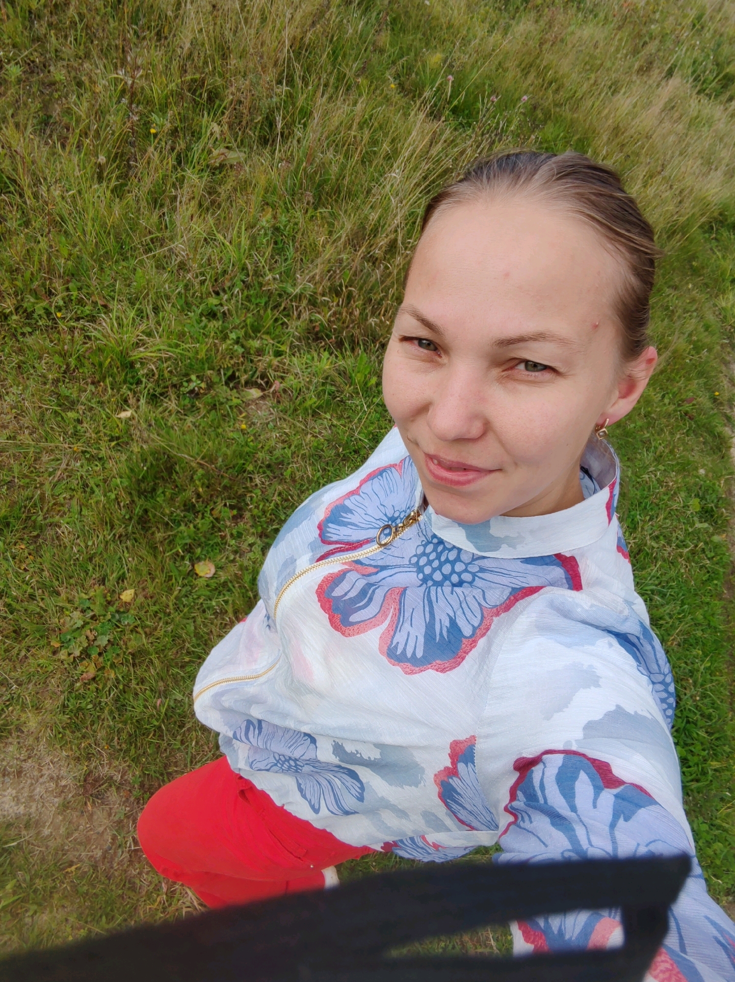 The image shows a young woman with brown hair pulled back and a light complexion, looking up at the camera. She is wearing a white hooded jacket with a blue and red floral pattern and red pants. The woman is outdoors, standing in a grassy area with some small yellow and pink flowers visible. Part of what appears to be a black object, possibly a camera or phone, is visible in the foreground, obscuring the bottom of the image. The background consists of tall, dry grass and some greener patches. The overall impression is a casual outdoor portrait. The location of Чухлэм, Russia, is not discernible from the image itself.