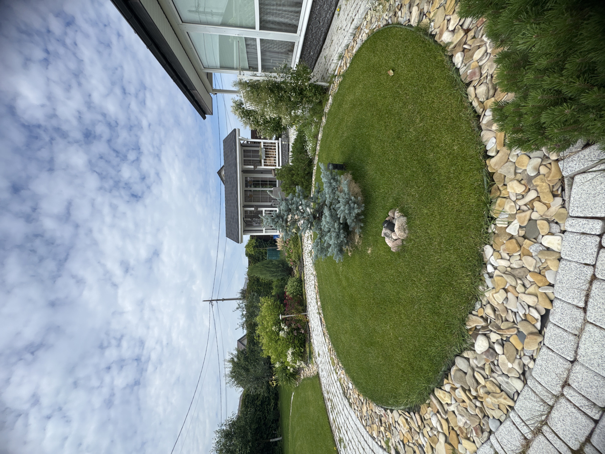 The image captures a well-maintained garden scene with a circular lawn as the focal point. A small blue spruce tree stands in the center of the lawn, surrounded by a ring of small rocks. The lawn is bordered by a pathway constructed of gray pavers and decorative pebbles in various shades of beige and yellow. To the right of the lawn, a section of a modern house with large windows and a dark roof is visible. In the background, a small, rustic-looking shed or outbuilding with a porch and windows is nestled amongst lush green foliage and trees. The sky above is a mix of blue and white clouds. The environment appears to be a suburban or rural residential area, given the presence of houses, gardens, and overhead power lines. The overall impression is one of a peaceful and aesthetically pleasing outdoor space. There are no people visible in the image.