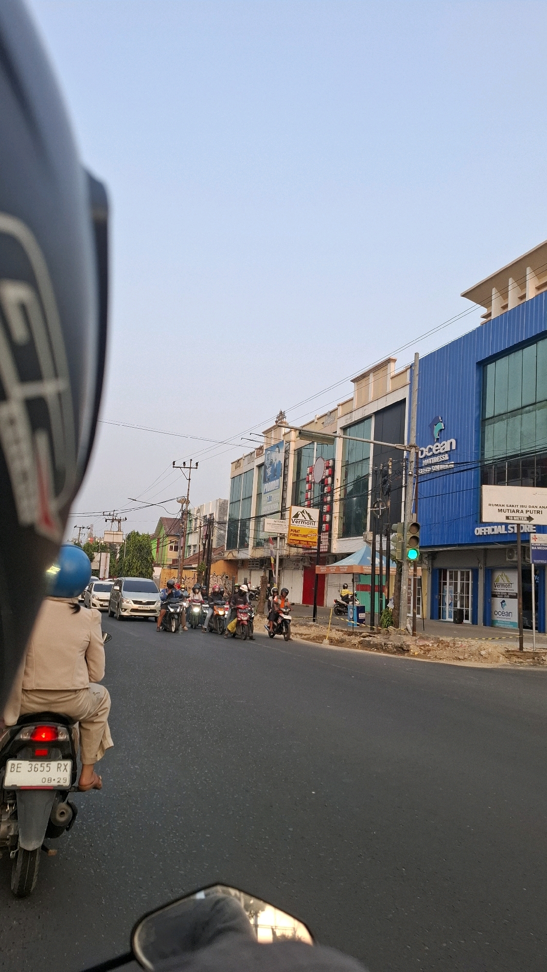 A first-person perspective shot captures a bustling street scene in Bandar Lampung, Indonesia, likely along Jalan Raden Intan. The camera is positioned behind a rider on a motorcycle, with the rider's helmet and back visible in the foreground. Ahead, a line of motorcycles and a silver car navigate the asphalt road, indicating moderate traffic. Several buildings line the street, featuring signage for businesses like "Ocean Mattress" and "Vermont Furniture." One prominent blue building displays the "Ocean" logo. A traffic light is visible and appears to be green. The sky is clear and pale blue, suggesting it is daytime. The overall atmosphere is one of everyday urban activity.