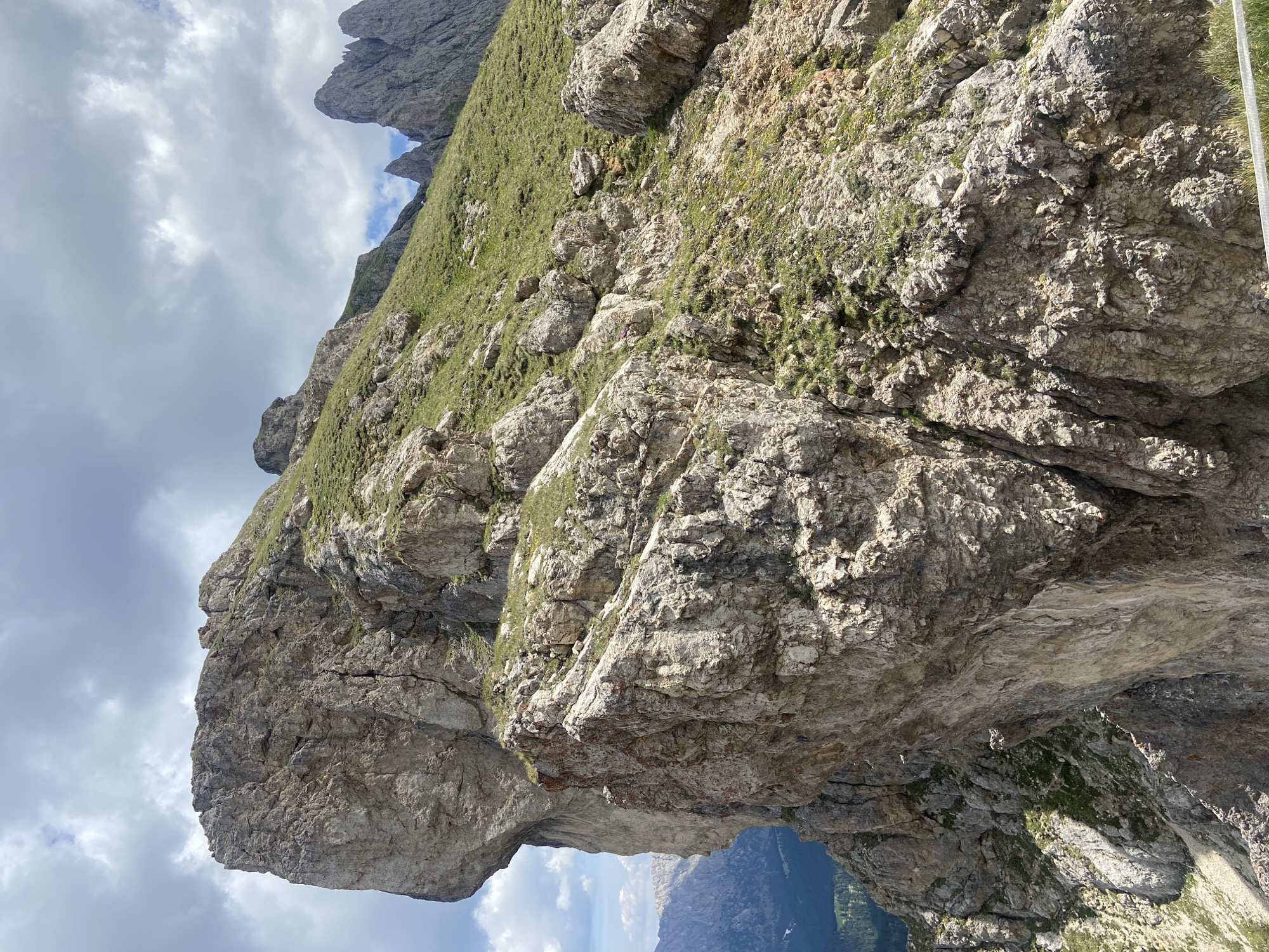 The image shows a dramatic mountain landscape in the Dolomites, Italy. In the foreground, a massive, sheer rock face dominates the left side of the frame, characterized by its rough texture and layered appearance. To the right and extending into the background, a steep, grassy slope dotted with rocks ascends towards a ridgeline. The sky above is partly cloudy, with a mix of white and grey clouds against a pale blue backdrop. In the far distance, other jagged mountain peaks are visible, suggesting a vast mountain range. The scene is natural and unpopulated, emphasizing the rugged beauty of the alpine environment.