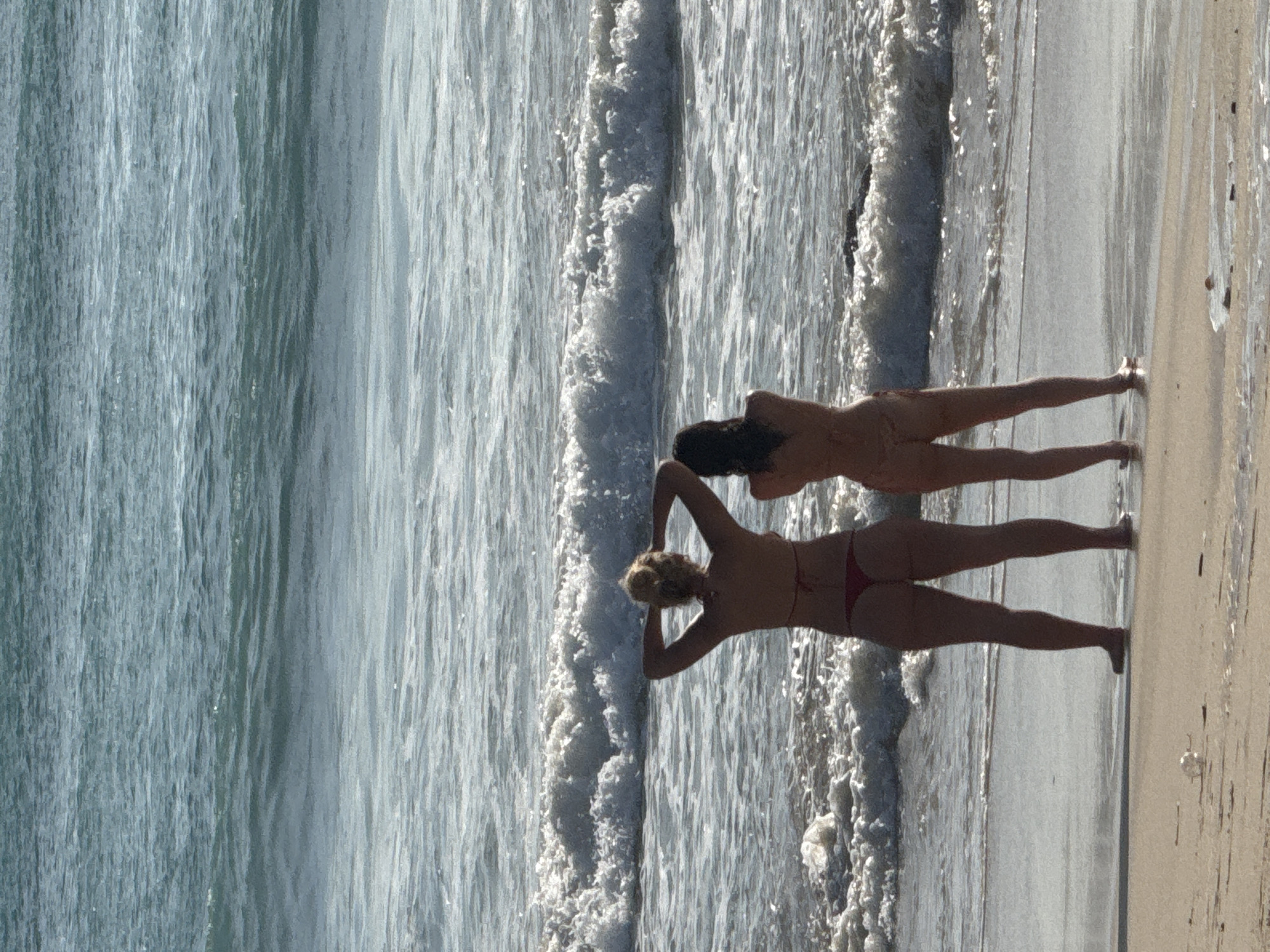 This image captures a bright, sunny beach scene in Kuta Selatan, Indonesia, featuring two individuals standing at the water's edge. The photograph is oriented vertically, but depicts a landscape view of the ocean and a sandy shore.
The two subjects, likely women, are positioned with their backs to the camera, facing the expansive ocean. They are wearing two-piece swimsuits. The individual closer to the viewer has blonde hair, possibly tied up, and is clad in a red bikini. Her arms are raised above her head, with hands clasped behind her neck. Slightly further away, the second individual has dark hair and is wearing a lighter-colored bikini, perhaps tan or light orange, and strikes a similar pose with arms raised behind her head. Their figures are slightly silhouetted against the bright light reflecting off the water.
The setting consists of the vast ocean, occupying the majority of the left and central parts of the image, and a sandy beach on the right. Waves with prominent white foam are actively breaking and rolling onto the shore, indicating dynamic water conditions. The ocean water exhibits varying shades of blue and green under the bright sunlight. The sand appears light in color, with areas near the water line appearing wet and reflective.
The scene conveys a sense of leisure and enjoyment of the coastal environment. The time of day appears to be bright daylight, suggesting midday or early afternoon, given the strong illumination and lack of long shadows. There is no visible text in the image.