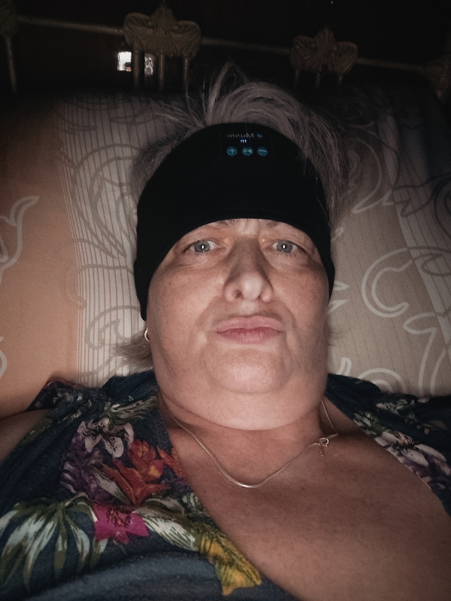 The image depicts an individual in a supine position, gazing directly at the camera in an indoor setting, specifically a bedroom in Krasnodar, Russia. The subject, an adult with light-colored, disheveled hair, possesses fair skin with visible freckles and light-colored eyes. They are wearing a black sleep headband equipped with illuminated blue controls for music playback (volume up/down, play/pause) and the mirrored text "music." The individual is attired in a dark top featuring a vivid floral pattern with elements of pink, purple, orange, green, and white. A thin, silver-colored snake chain necklace with a small clasp or pendant rests on the collarbone, and a small gold-colored hoop earring is visible in the right ear. The subject is resting on a light-colored pillow adorned with a subtle, repeating pattern of swirling lines. The background above the pillow is dark, revealing parts of an ornate, dark-colored headboard or bed frame with decorative elements. A small, bright rectangular light source is faintly visible in the upper left portion of the background. The scene is dimly lit, consistent with an evening or nighttime environment.