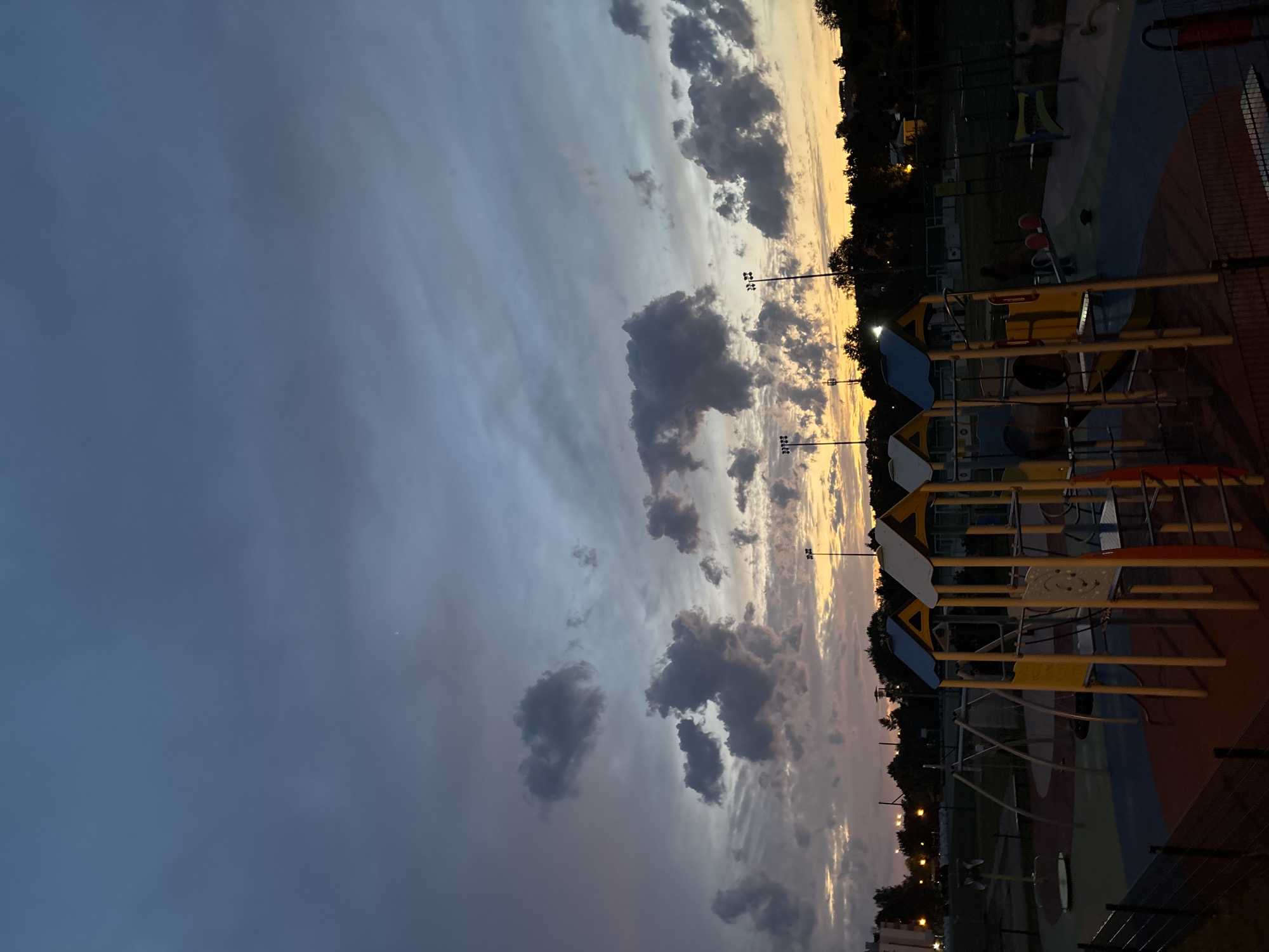 This is a nighttime outdoor photograph of a playground at dusk. The sky is filled with dramatic clouds in shades of dark blue, gray, and hints of orange and yellow from the setting sun on the horizon. In the foreground, a colorful playground structure with yellow and blue accents is visible, along with swings and a merry-go-round. The ground is a safety surface in various colors, including red, blue, and green. Tall light poles with floodlights are scattered throughout the park. In the background, trees and buildings are silhouetted against the twilight sky. The overall impression is one of a peaceful and serene evening scene. This playground is located in Lublin, Poland, specifically in the LSM (Lubelska Spółdzielnia Mieszkaniowa) district, known for its residential areas and green spaces.