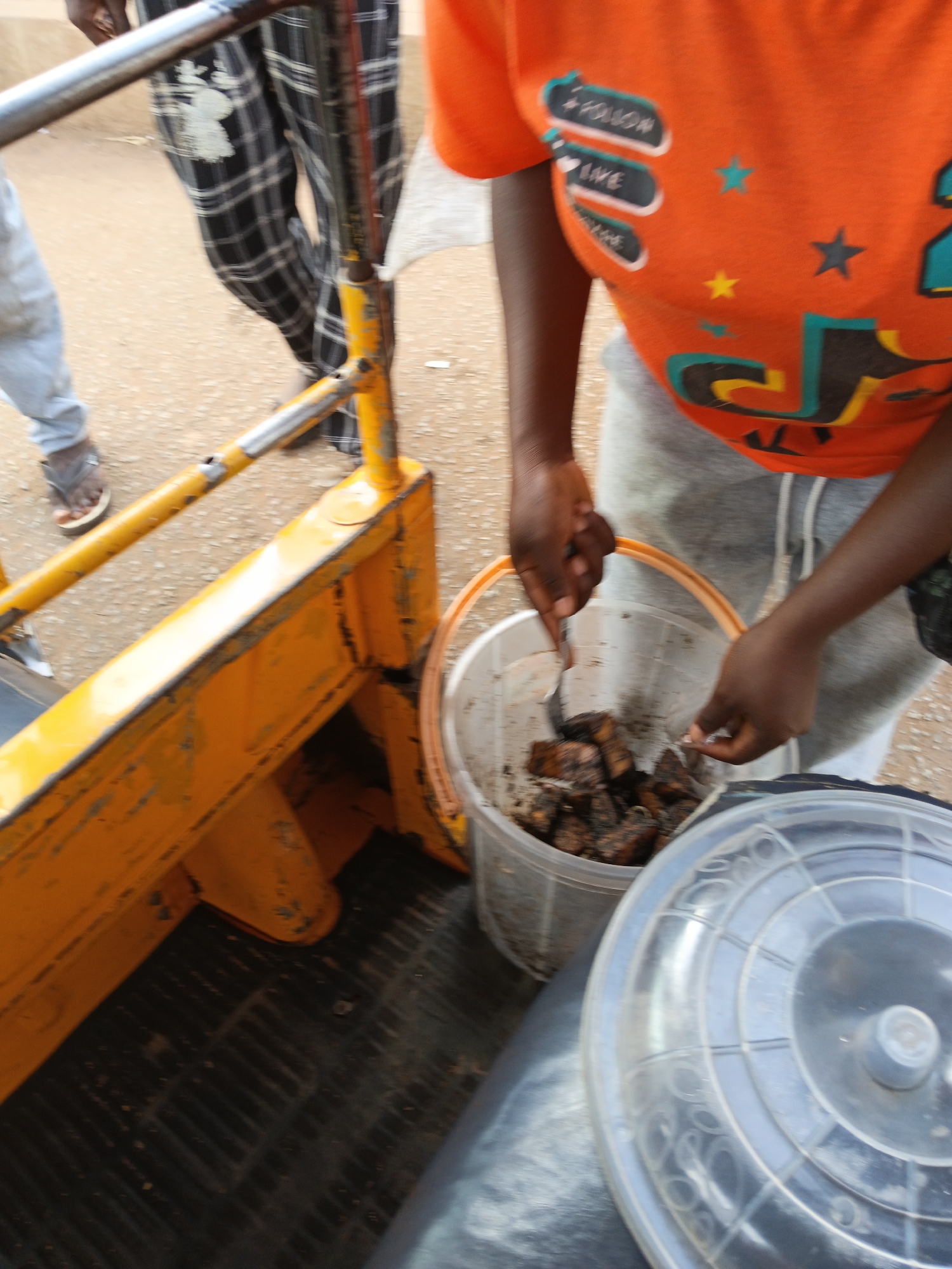 In an outdoor environment, likely a street, a person wearing a bright orange t-shirt with a social media-themed graphic is interacting with a plastic bucket filled with what appears to be grilled or fried food. The person's hands are visible, and they are holding a fork or utensil, suggesting they are either serving or sampling the food. The bucket is transparent, allowing a clear view of the dark-colored food pieces. In the background, another person is wearing checkered pants and a white shirt, standing near a yellow metal structure that resembles part of a vehicle or cart. The ground is covered with dirt and loose gravel. A clear plastic lid is placed on a black surface next to the food bucket. The overall scene suggests a casual street food vendor or someone preparing food outdoors.
