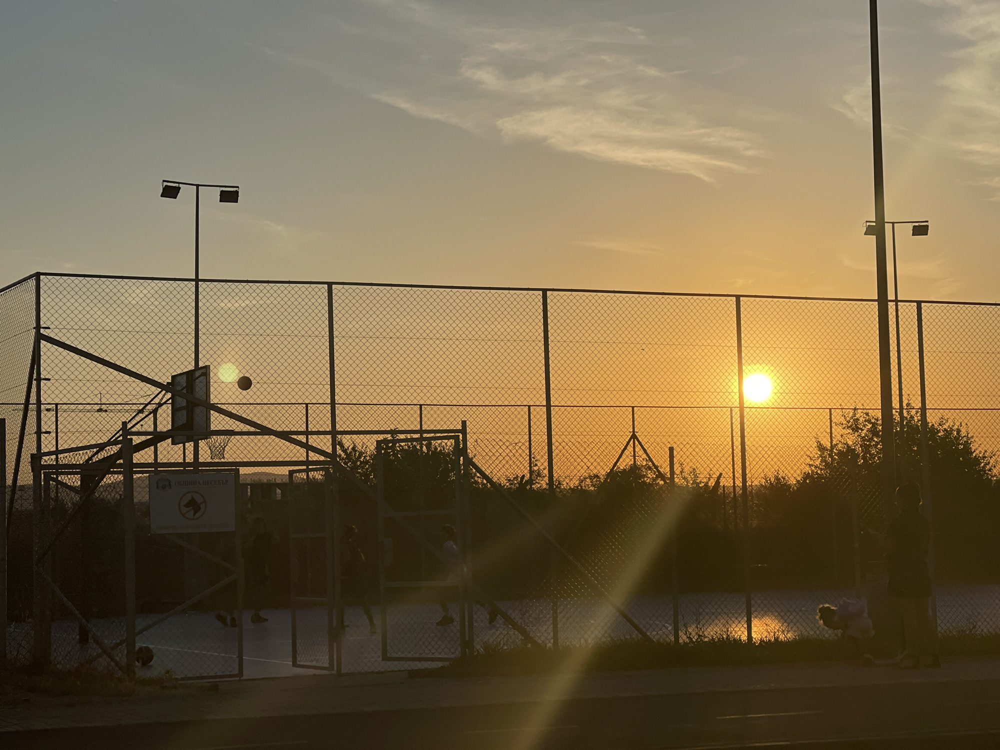 This image captures a sunset over a basketball court located in Nesebar, Bulgaria. The sun, a bright orb, is visible in the background, casting a warm, orange glow across the sky and a chain-link fence that encloses the court. Silhouetted figures of people can be seen on and around the court, some appearing to be playing basketball. The court itself is dimly lit by the setting sun, with light poles also visible. Vegetation and distant structures are faintly discernible beyond the fence. The scene is characterized by the golden hour light, creating a serene and atmospheric ambiance.