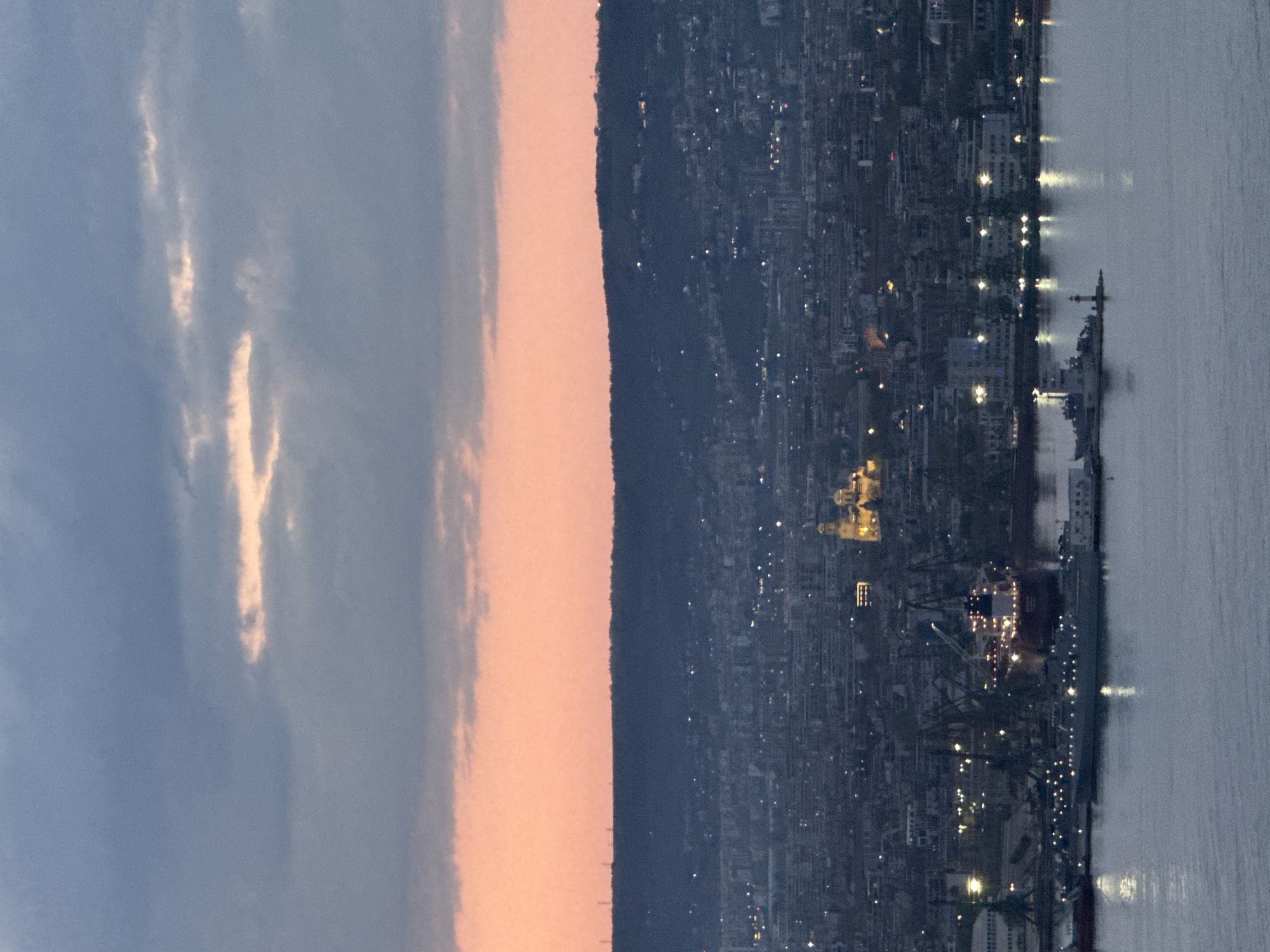 This panoramic image depicts the city of Varna, Bulgaria, at dusk or dawn, showcasing a captivating transition between day and night. The scene is structured horizontally, from top to bottom, revealing the sky, a mountainous backdrop, and the illuminated city leading to a body of water.
The upper portion of the frame features a sky transitioning from a pale blue-grey on the left, where delicate, elongated clouds catch a soft, ethereal light, to a warm peach and orange glow along the horizon to the right.
Below the sky, a dark, densely forested mountain or hill range forms a prominent silhouette, stretching across the middle of the image and separating the atmospheric sky from the urban sprawl.
The lower half of the scene presents the city of Varna, illuminated by countless artificial lights. Buildings, both residential and commercial, twinkle against the deepening twilight. A distinctive landmark, likely the Dormition of the Theotokos Cathedral, stands out with its bright yellow/golden domes and illuminated facade, situated prominently within the cityscape. Adjacent to the city, a bustling port area is visible, featuring various industrial structures, including cranes.
The lowest section of the image is dominated by a calm body of water, likely the Black Sea or Varna Bay. The water reflects the city lights and those from several vessels docked in the harbor. A large ship, with its decks brightly lit, is a notable presence, alongside other boats and what appears to be a lighthouse or navigation beacon.
The overall mood is serene and peaceful, capturing the quiet beauty of a city as it transitions between day and night. No specific human activity is discernible, and no text is visible in the image.